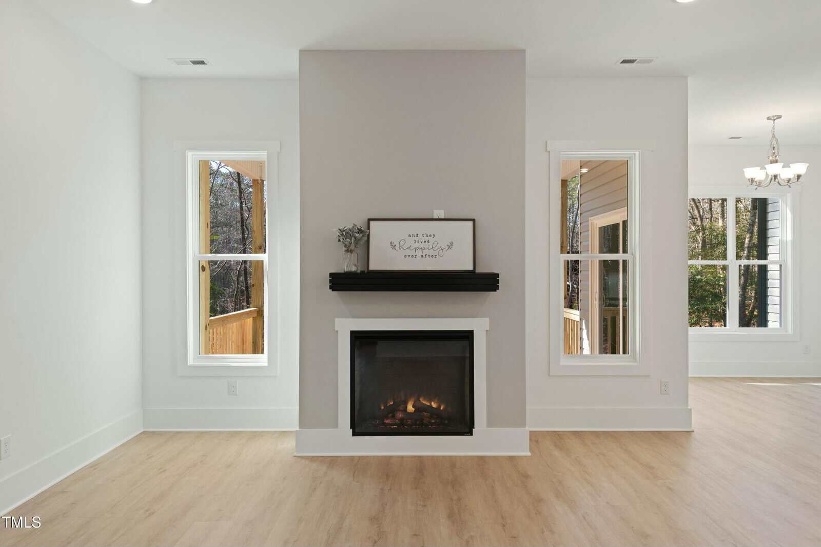 A modern living room with light wood flooring, a white fireplace centered on a white wall, two tall windows on either side, and a small framed sign and vase on the black mantel. A dining area is visible on the right.