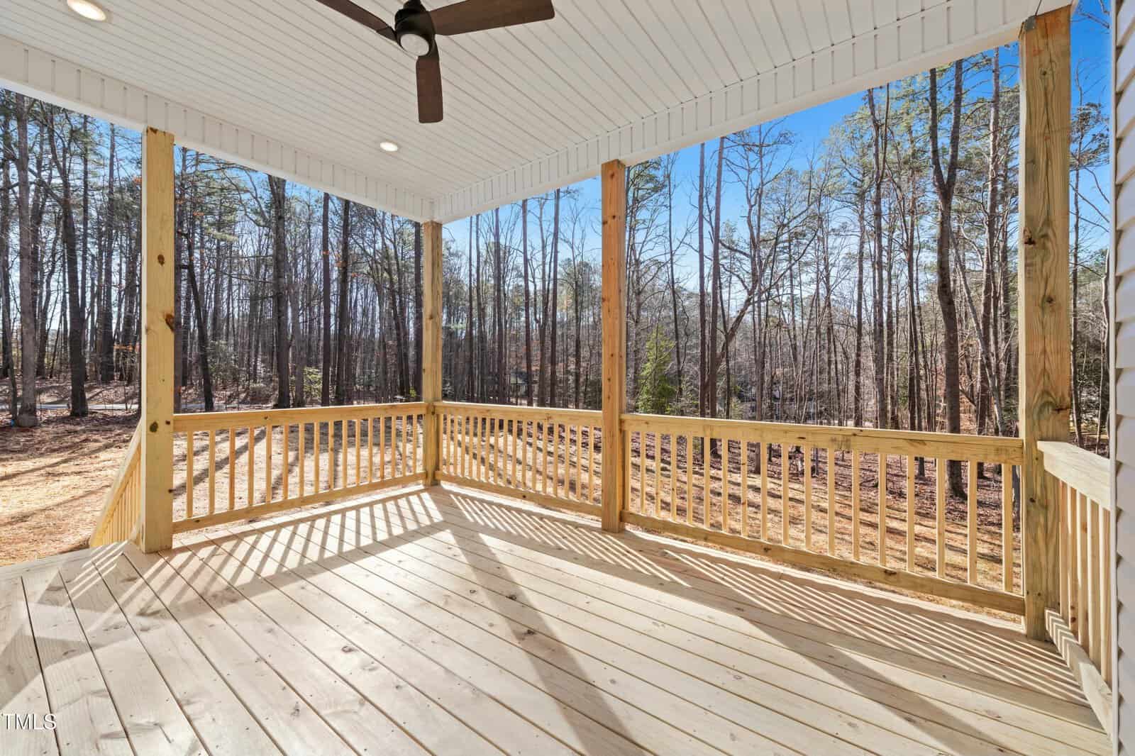 A covered wooden deck with a ceiling fan and railing overlooks a wooded area with tall trees and clear blue sky. Sunlight casts shadows on the deck floor.