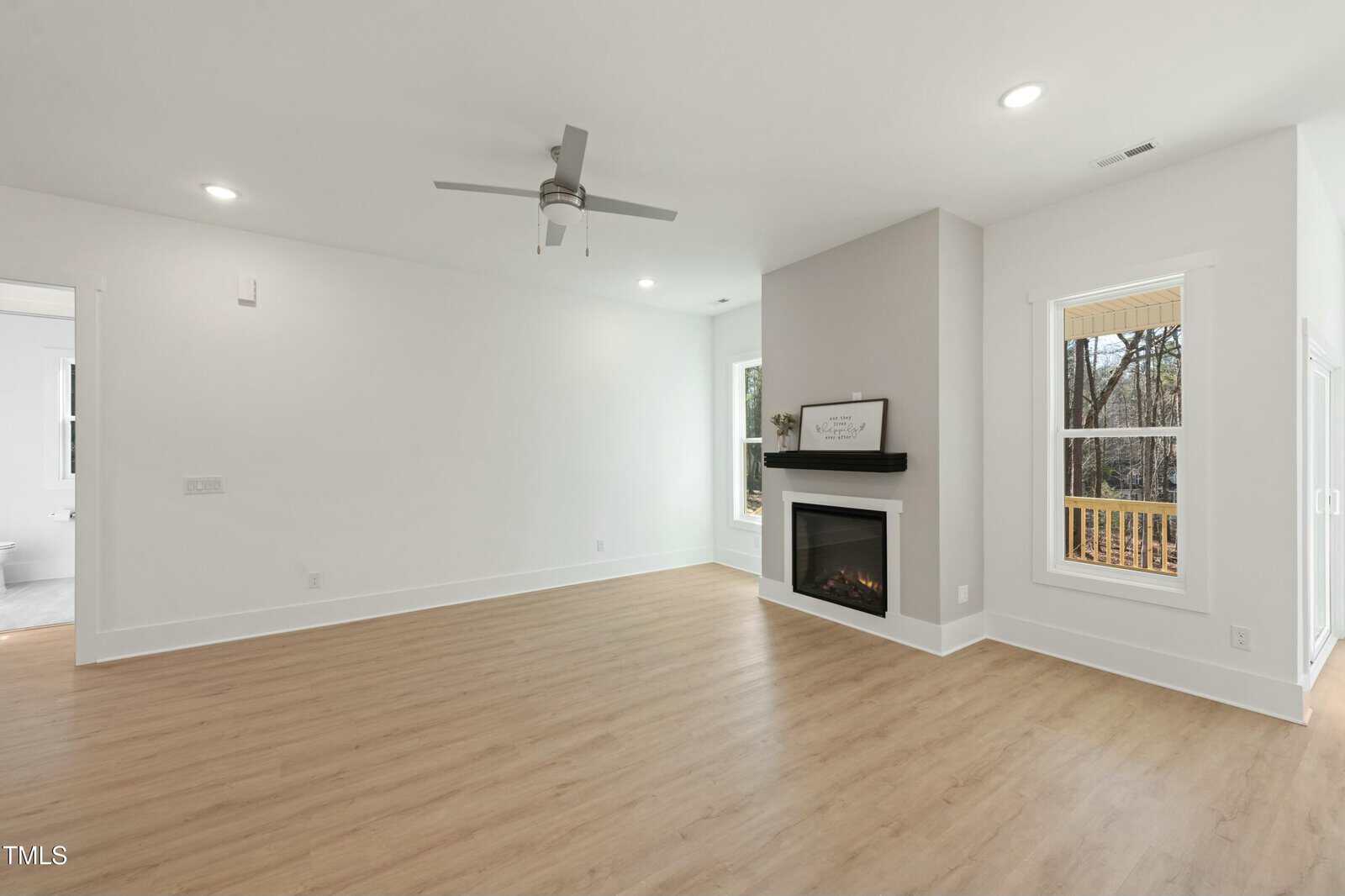 A bright, empty living room with light wood floors, white walls, a ceiling fan, a modern corner fireplace, and large windows showing a deck and trees outside.