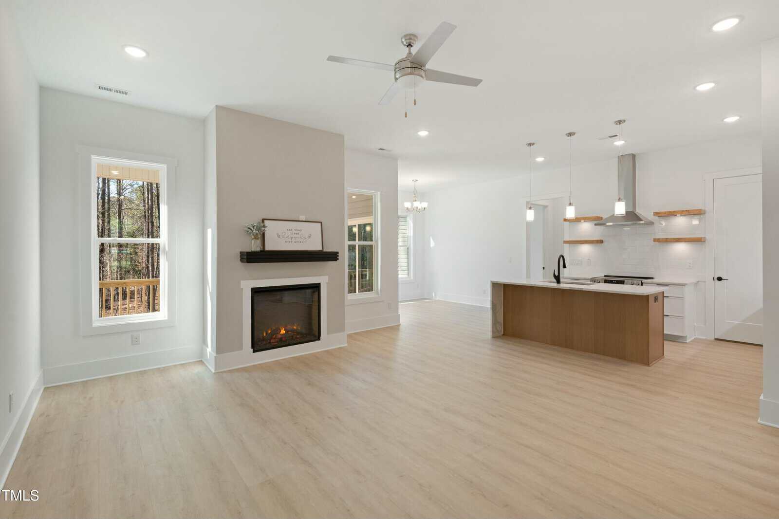 Open-concept living area with light wood floors, a modern fireplace, large windows, and a kitchen featuring a wood island, pendant lights, white cabinets, and stainless steel appliances. White walls and ceiling fans are visible.