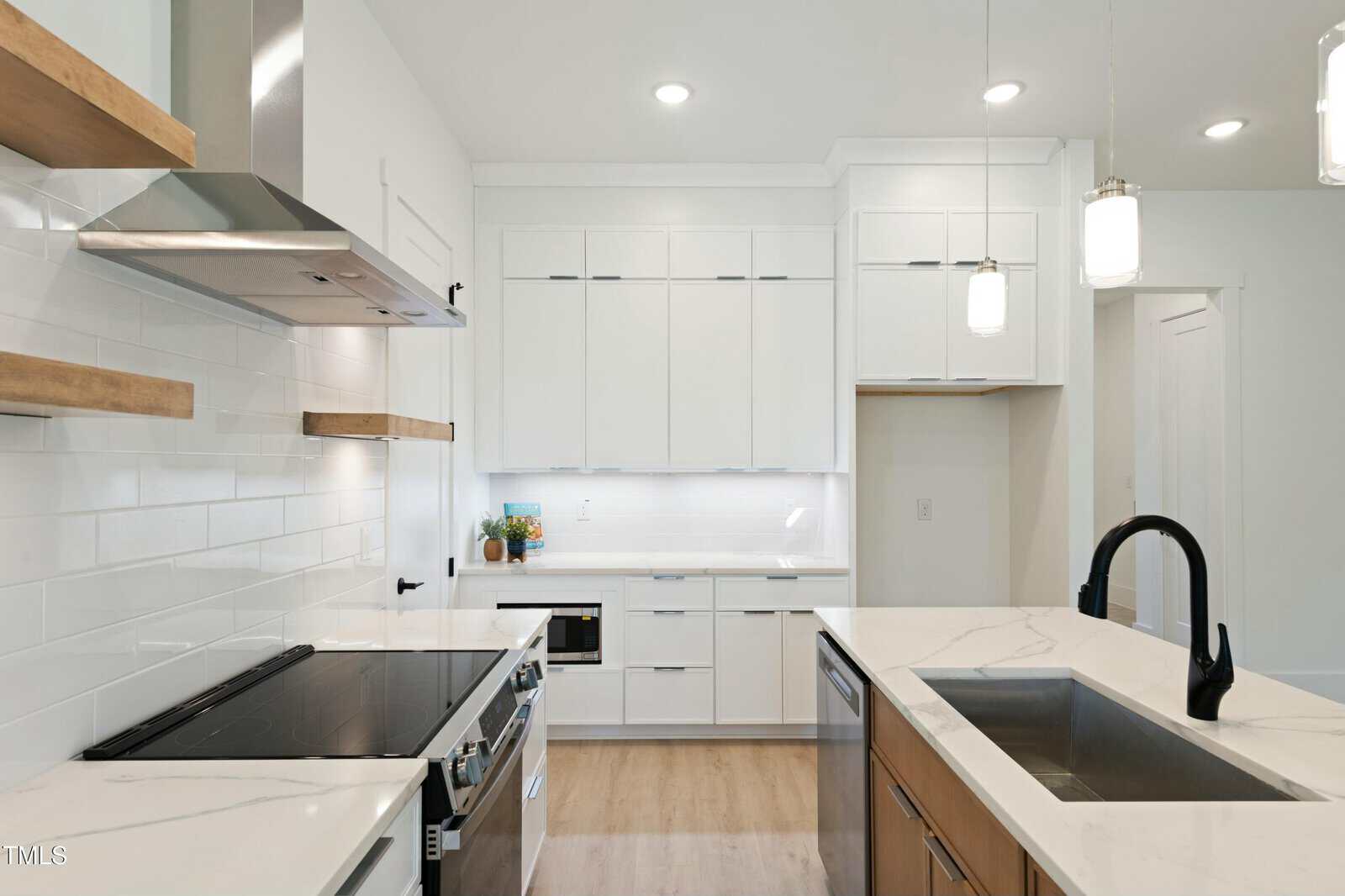 Modern kitchen with stainless steel appliances, white cabinets, a black faucet, marble countertops, wooden shelves, white subway tile backsplash, and pendant lights over the island. Light wood flooring is visible.