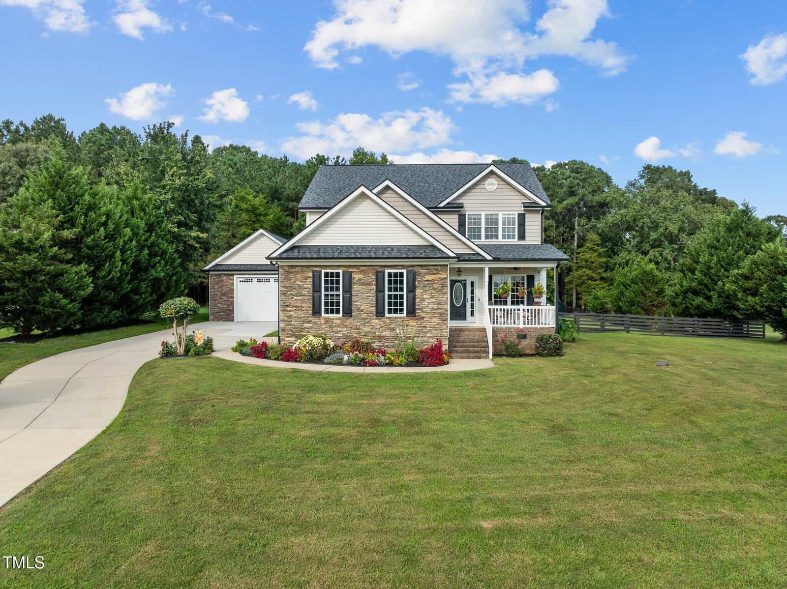 A suburban two-story house at 1052 Bluebell Lane in Wake Forest boasts a brick and siding exterior with a charming front porch. The property features a manicured lawn, curved driveway, and is surrounded by trees and a wooden fence under a partly cloudy sky.