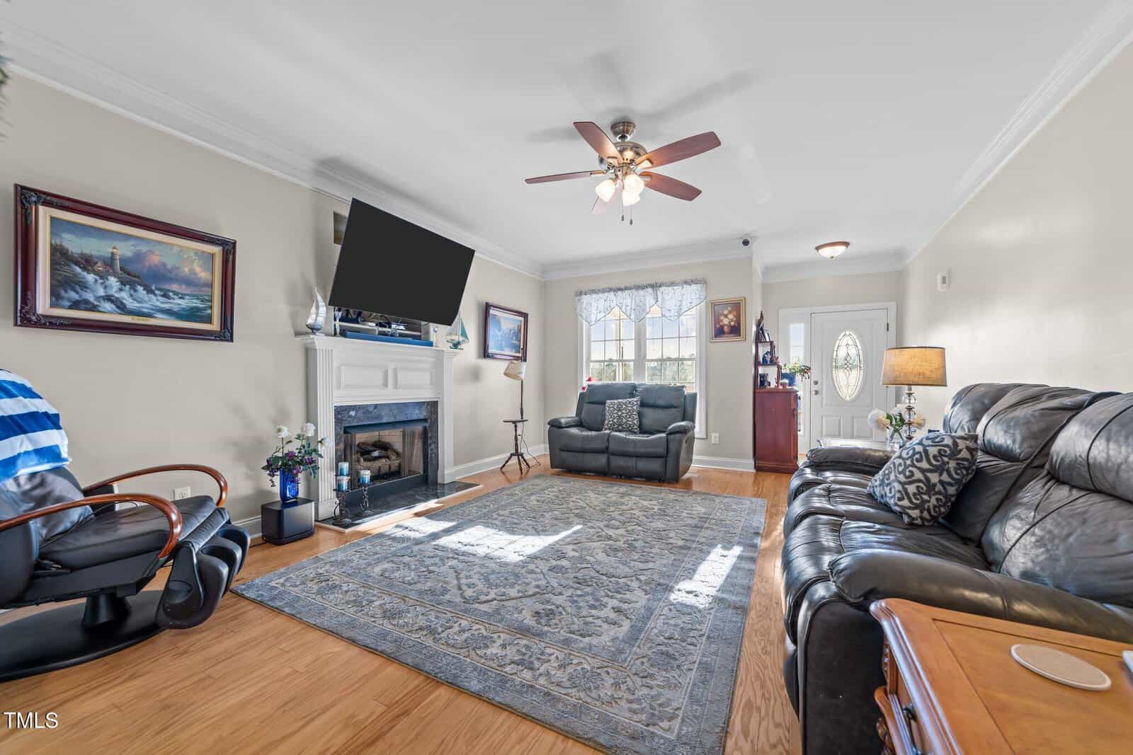 A living room features a leather recliner, loveseat, and chair. A TV is mounted above a fireplace. A ceiling fan hangs overhead, with framed art and a window on the walls. A patterned rug covers the wooden floor. A door is visible in the background.