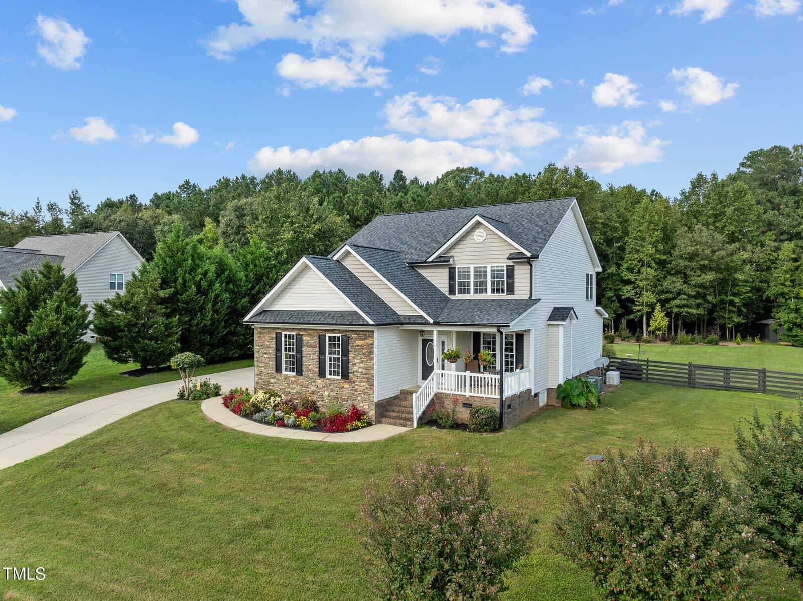 A two-story house with a brick and siding exterior features a front porch and landscaped yard. A driveway leads to the house, and it is surrounded by grass and trees under a partly cloudy sky.