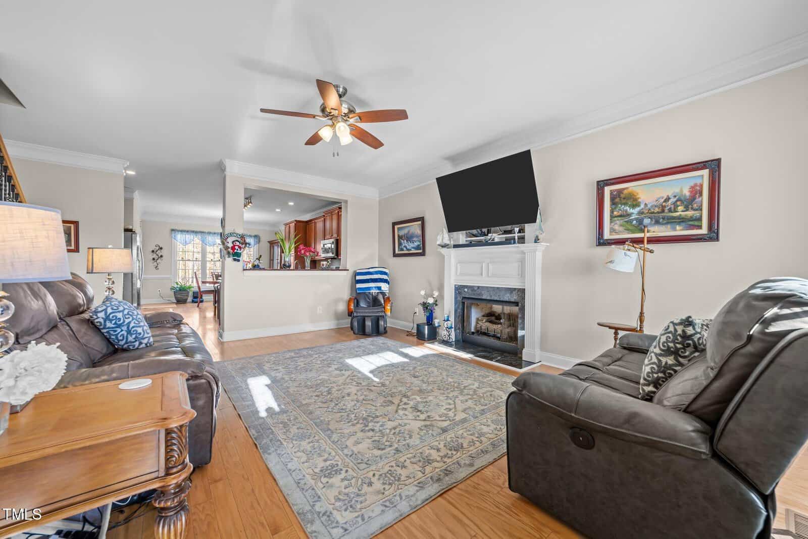 A cozy living room with a mounted TV above a white fireplace, surrounded by a gray sofa and armchair. A patterned rug covers the wooden floor. Paintings and lamps adorn the room, and a ceiling fan is above. The kitchen is visible in the background.