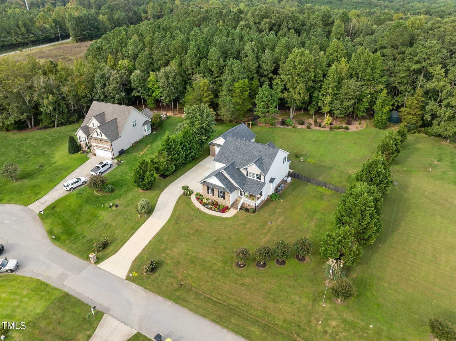 An aerial view of a suburban neighborhood featuring three houses surrounded by grass and trees. The central house has a light exterior and a gray roof, with a curved driveway. Dense forest is visible in the background.