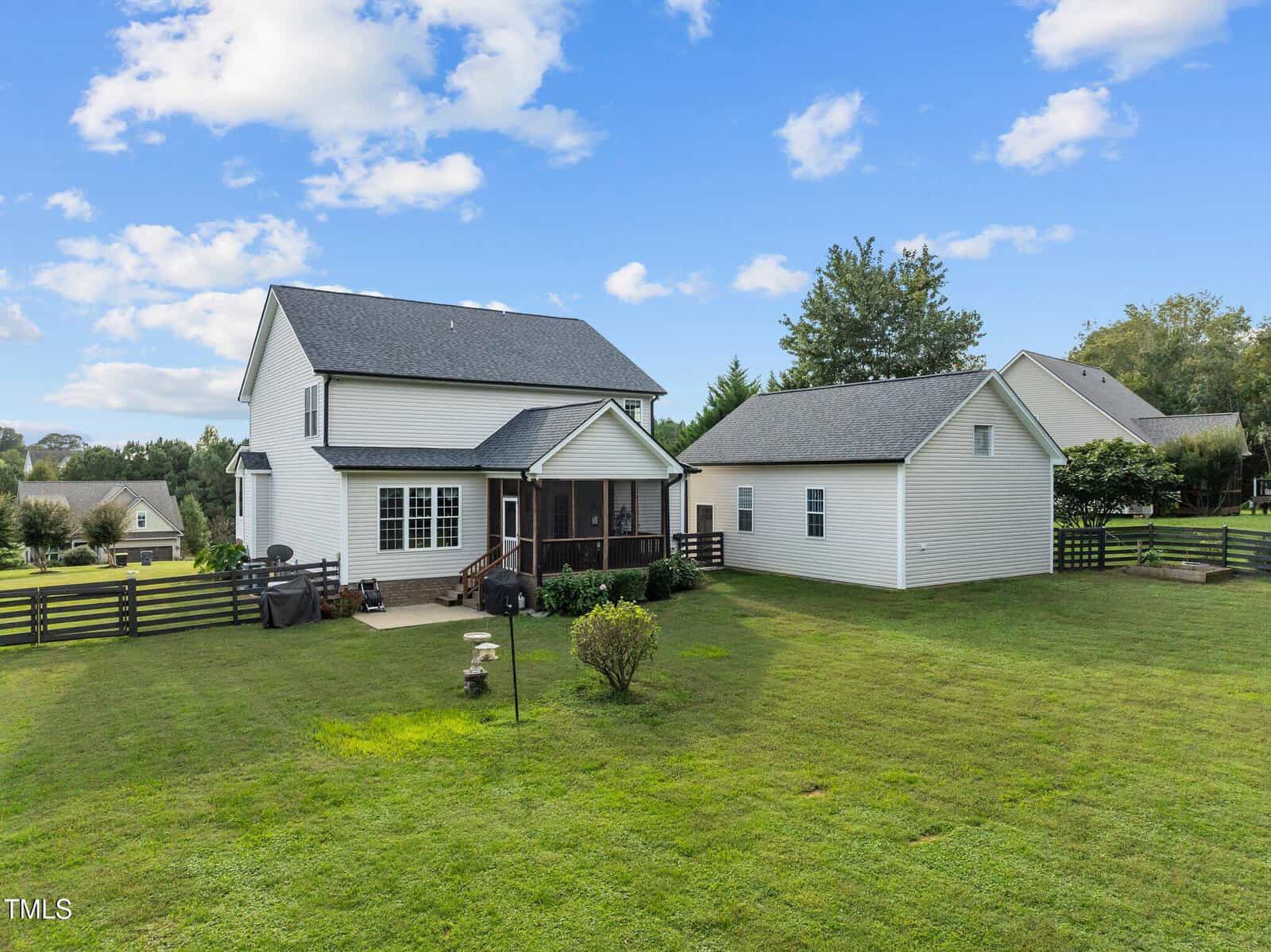 Two-story white house with a dark roof, featuring a screened porch and an adjacent detached garage. The house is surrounded by a spacious green lawn with a few shrubs and a wooden fence, set against a partially cloudy blue sky.