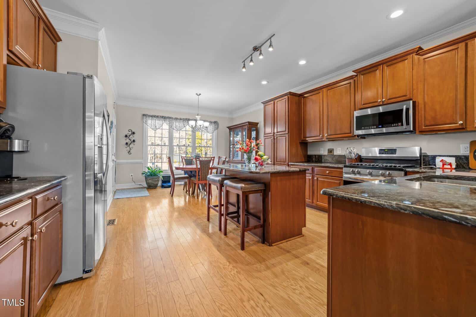 Spacious kitchen with wooden cabinets and a center island featuring granite countertops. Stainless steel appliances are present, along with a dining table in an adjoining area. A large window allows natural light to fill the room.