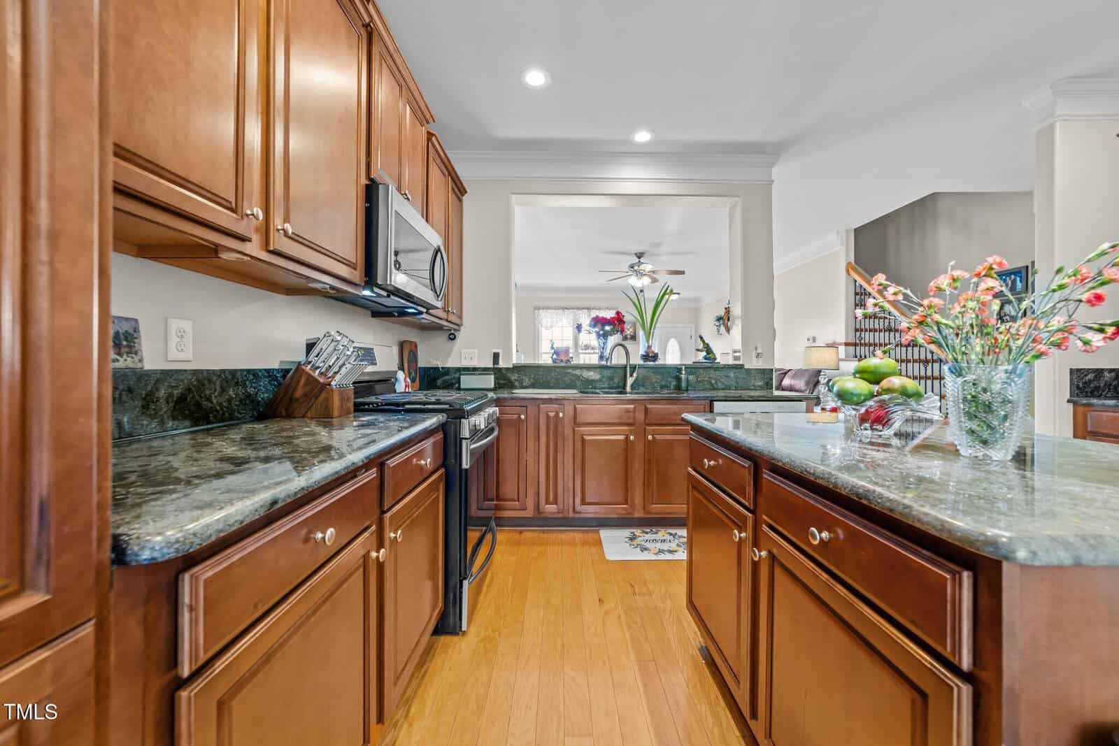 A spacious kitchen with wooden cabinets and granite countertops. A stainless steel microwave is built into the cabinetry. There's a vase with flowers on the counter and a bowl of fruits. The kitchen opens to a living area with a ceiling fan.