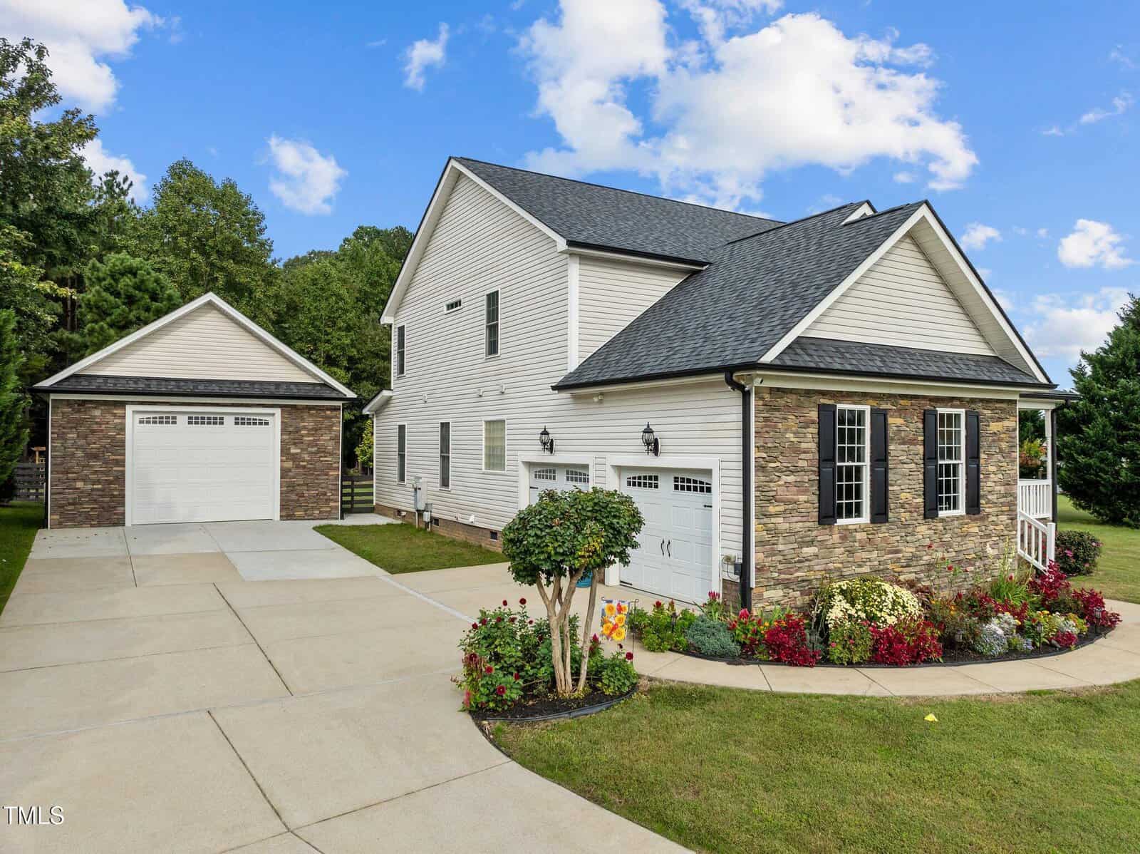 A two-story house with white siding and stone accents, featuring black shutters and a gray shingled roof. The front yard has landscaped bushes and flowers. A concrete driveway leads to a detached garage matching the house's exterior.