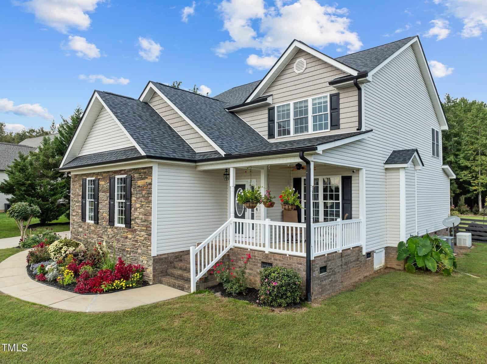 A two-story house with a mix of stone and siding exterior. It has a covered porch with a hanging flower basket. The front yard features a curved walkway and colorful flower beds. The sky is blue with scattered clouds.