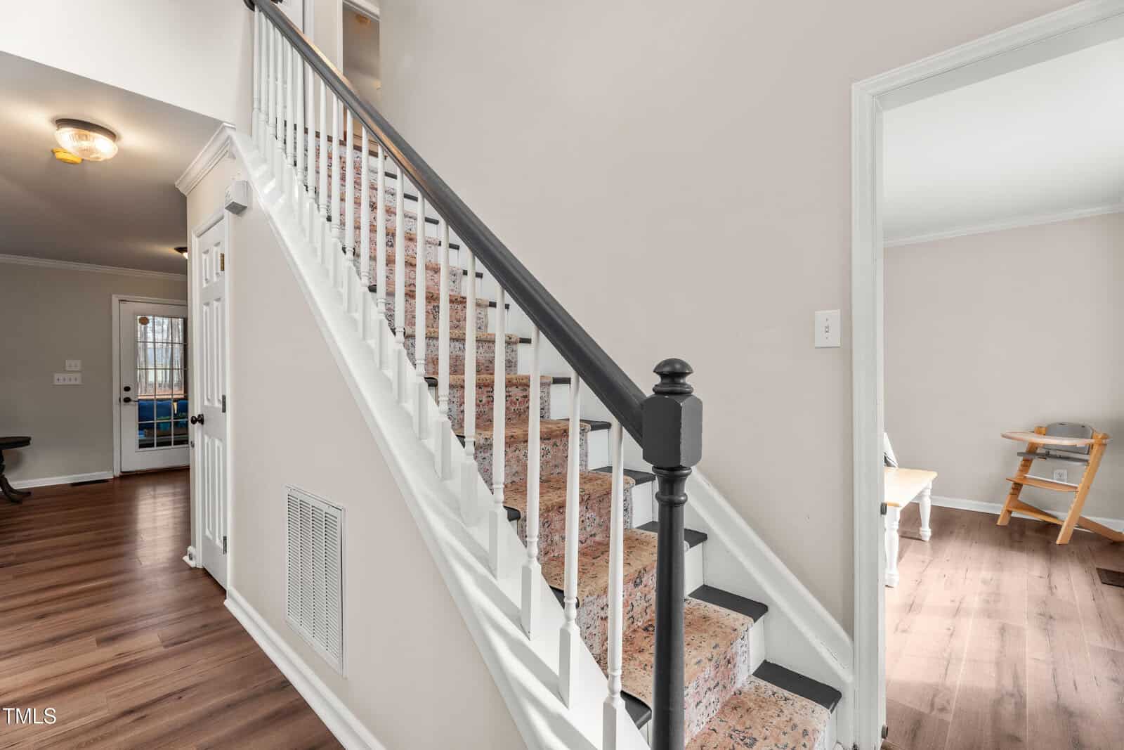 A staircase with a black handrail and patterned carpet leads upward beside a hallway with wood flooring. An open doorway on the right shows part of a room with a wooden chair and table.