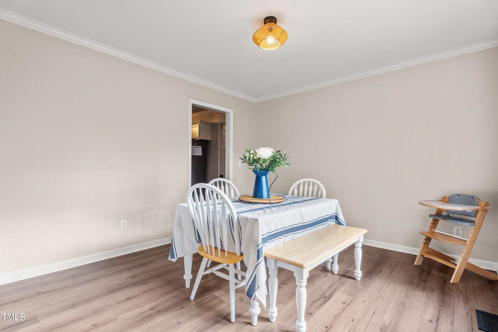A dining area with a table covered by a striped tablecloth, surrounded by two white chairs and a wooden bench. A vase with flowers sits on the table. There is a wooden high chair in the corner and a ceiling light above.