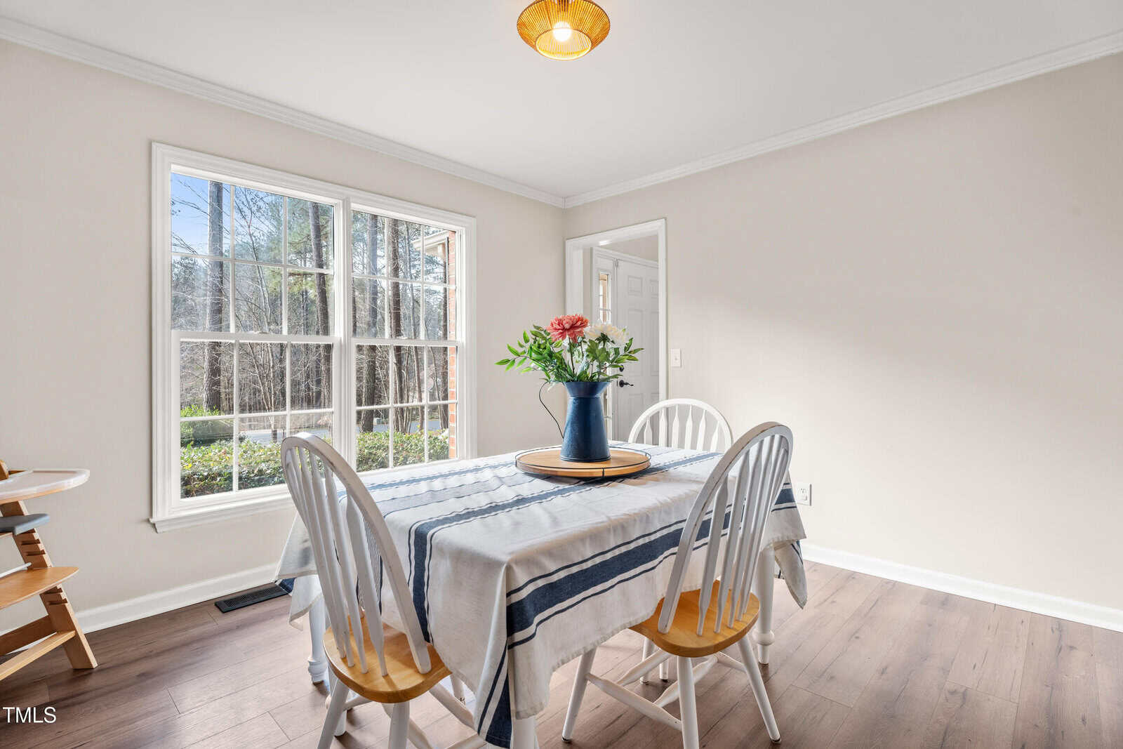 A bright dining room with a table covered by a striped tablecloth, four white chairs, a vase with flowers, large window letting in natural light, and light wood flooring.
