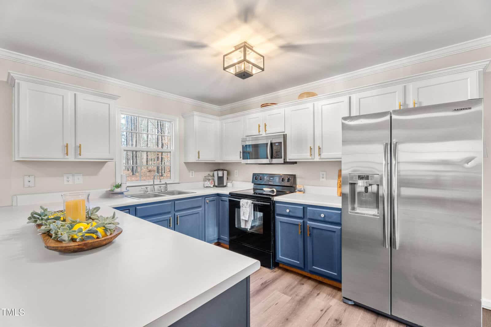 Modern kitchen with white upper cabinets, blue lower cabinets, stainless steel appliances, and white countertops; a bowl with decorative greenery and lemons sits on the counter; a window provides natural light.