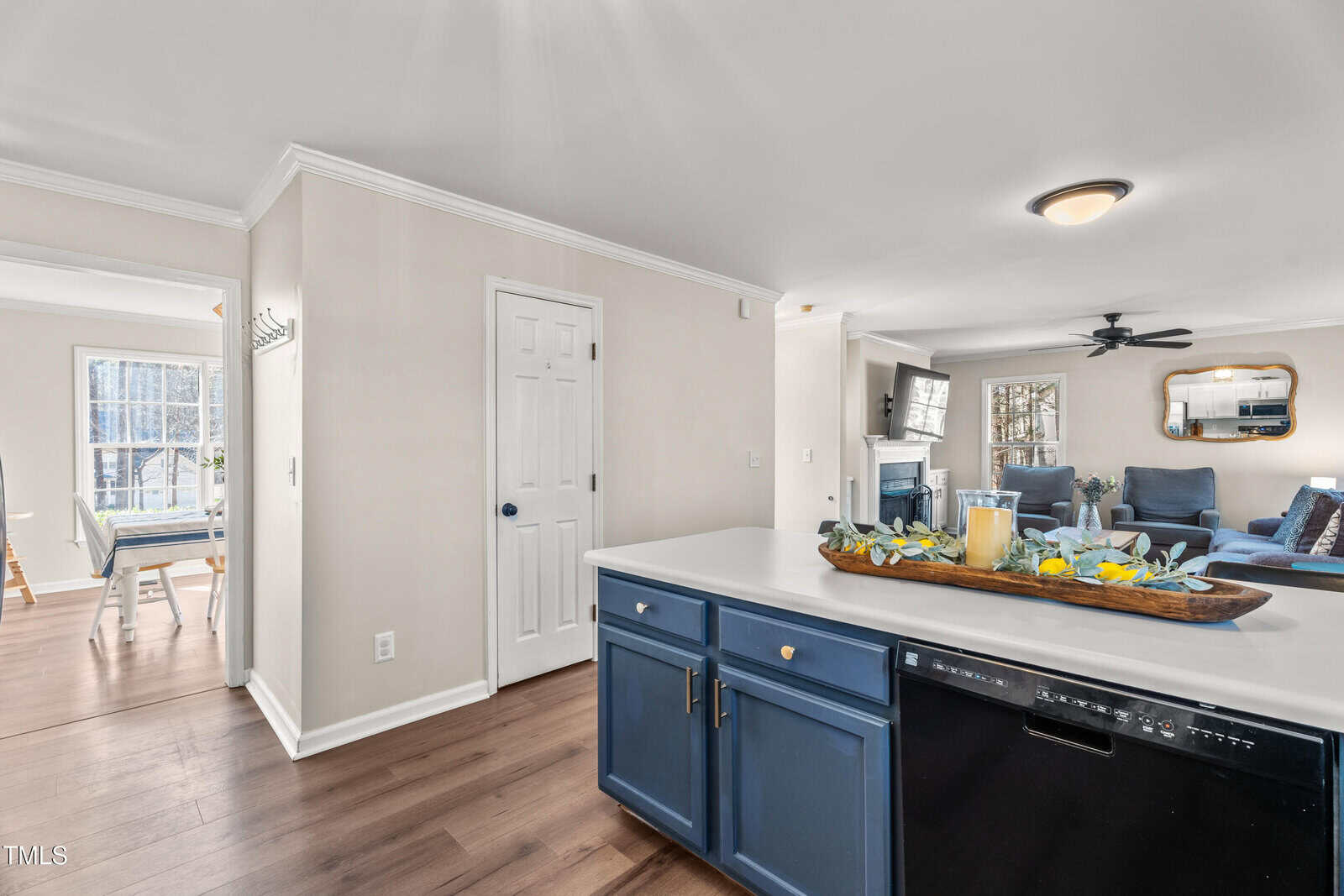 View from a kitchen island with blue cabinets and a dishwasher, showing a living room with seating, a wall-mounted TV, a mirror, and a glimpse of a dining area through an open doorway.