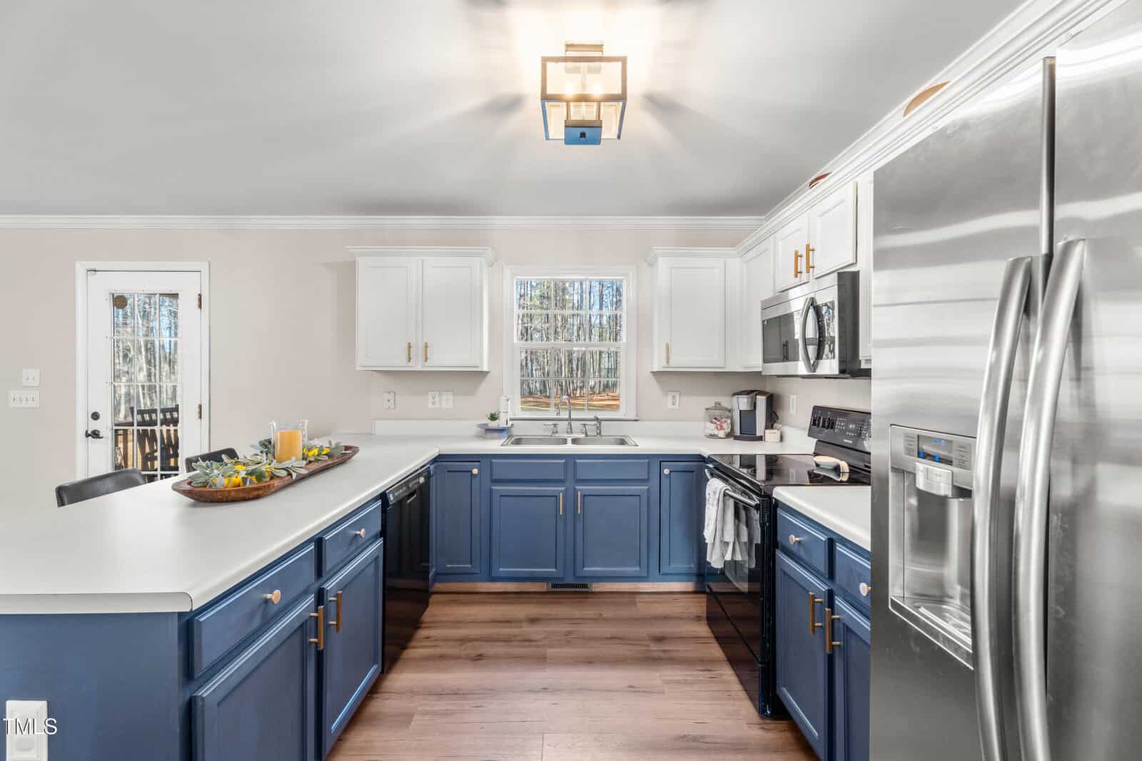 Modern kitchen with blue lower cabinets, white upper cabinets, stainless steel appliances, a central window, and a tray with juice and breakfast items on the counter. Light wood flooring and neutral walls.