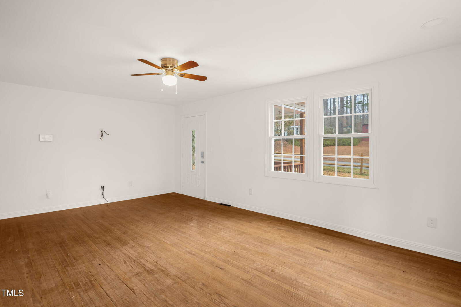 A sparse, empty room with white walls and hardwood flooring. A ceiling fan with a light is centered on the ceiling. Two windows let in natural light, and there is a visible door. Electrical outlets and a wall-mounted fixture are also present.
