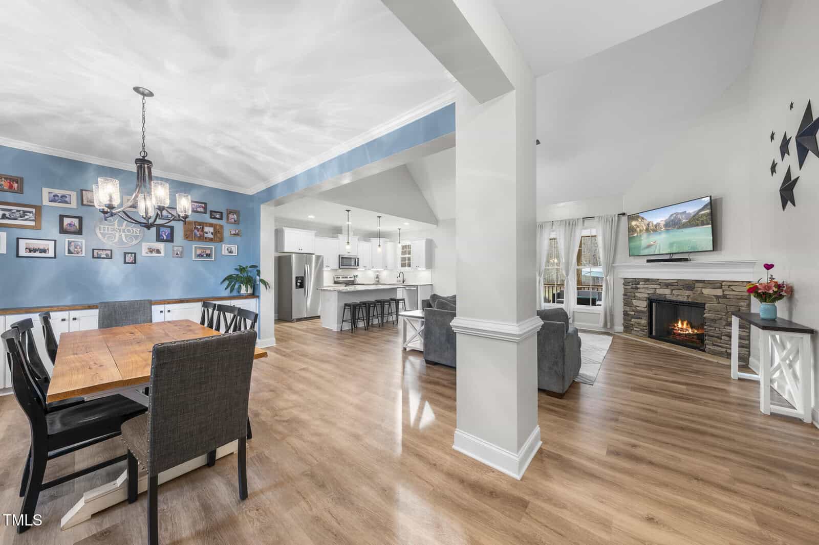 Open-concept living and dining area with wood flooring, blue accent wall with photo gallery, wooden dining table, black chairs, modern kitchen with white cabinetry, living room with fireplace and wall-mounted TV.