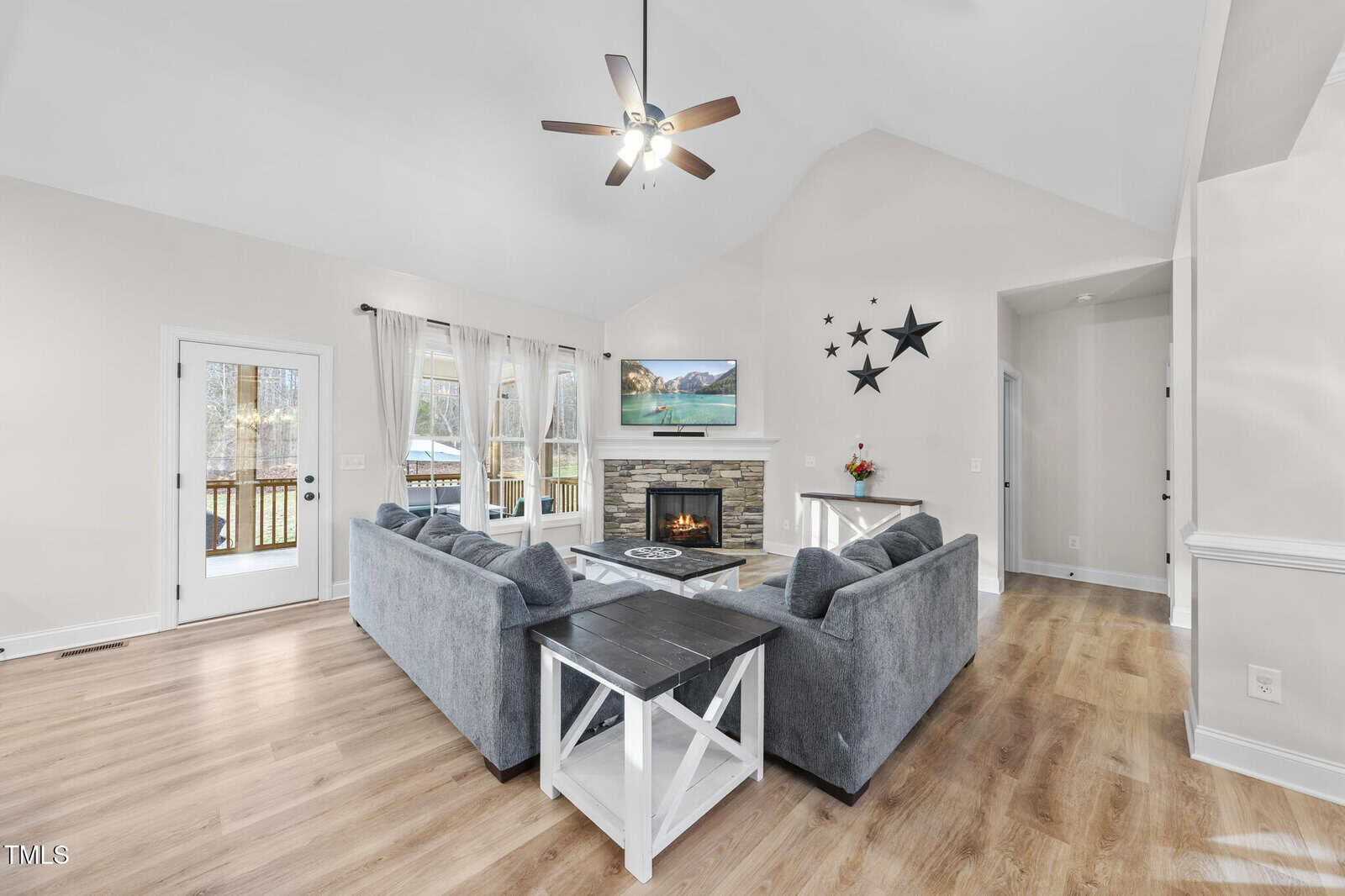 A living room with light walls, wood flooring, a gray sectional sofa, a fireplace with a TV above it, star decorations on the wall, and large windows with white curtains. A ceiling fan hangs above.
