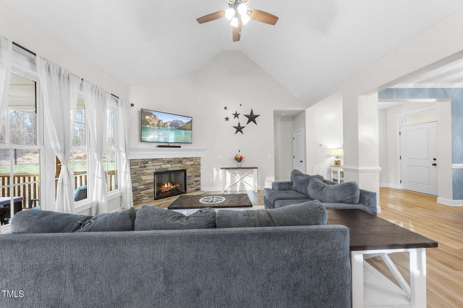 A bright living room with gray sofas, a wooden coffee table, a stone fireplace with a TV above it, star decorations on the wall, hardwood floors, and large windows with white curtains overlooking a porch.