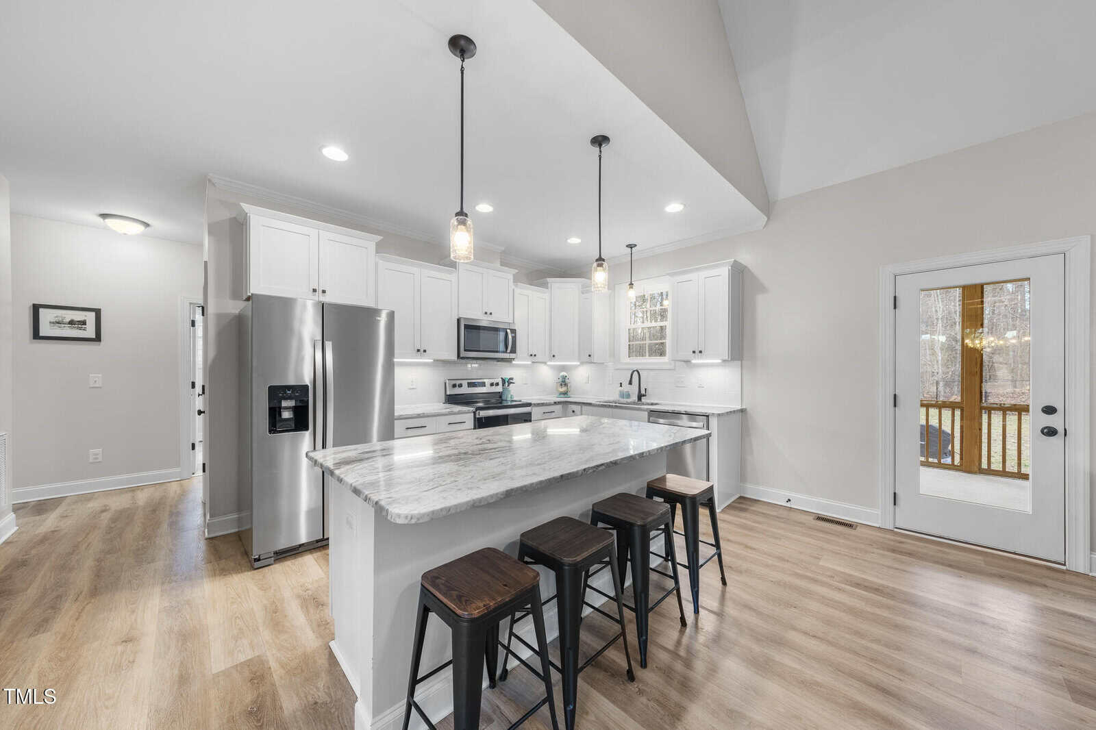 Modern kitchen with white cabinets, stainless steel appliances, a marble island countertop, three black stools, pendant lights, wood flooring, and a door leading outside. The space is bright with recessed ceiling lights.