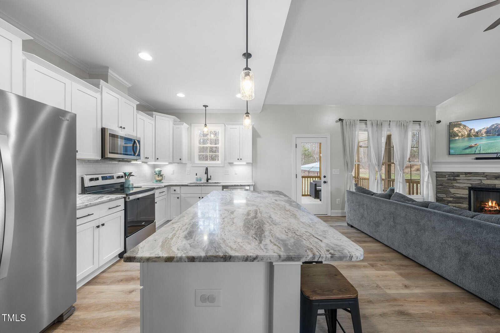 Modern kitchen with white cabinets, stainless steel appliances, marble island with stools, pendant lights, open living area with gray sectional sofa, fireplace, wall-mounted TV, large windows, and glass door leading outside.