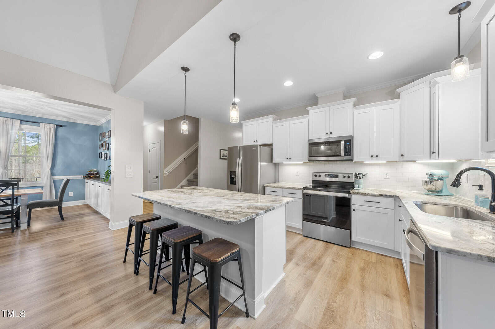 Modern kitchen with white cabinets, stainless steel appliances, a marble island with three stools, pendant lighting, wood flooring, and an adjoining dining area with a window and blue accent wall.