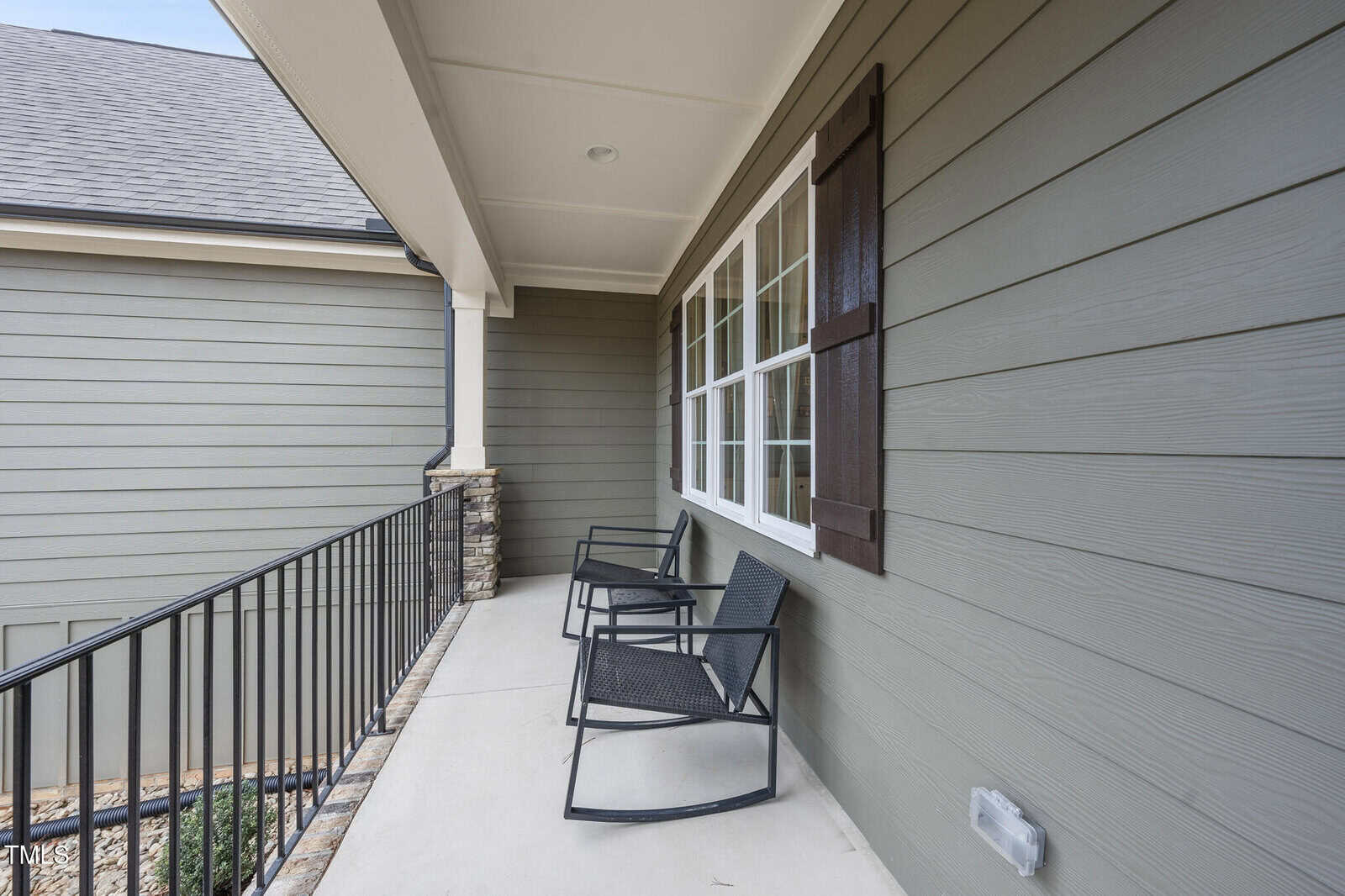 A covered porch with two black metal chairs, beige siding, white-framed windows, and a black metal railing. Stone accents are visible on the railing posts.
