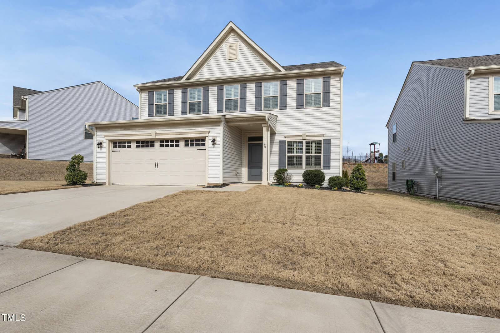 Two-story beige house with white trim and a double garage, situated on a plot with a brown, dry lawn. The house has a covered front porch and is flanked by similar neighboring homes under a clear blue sky.