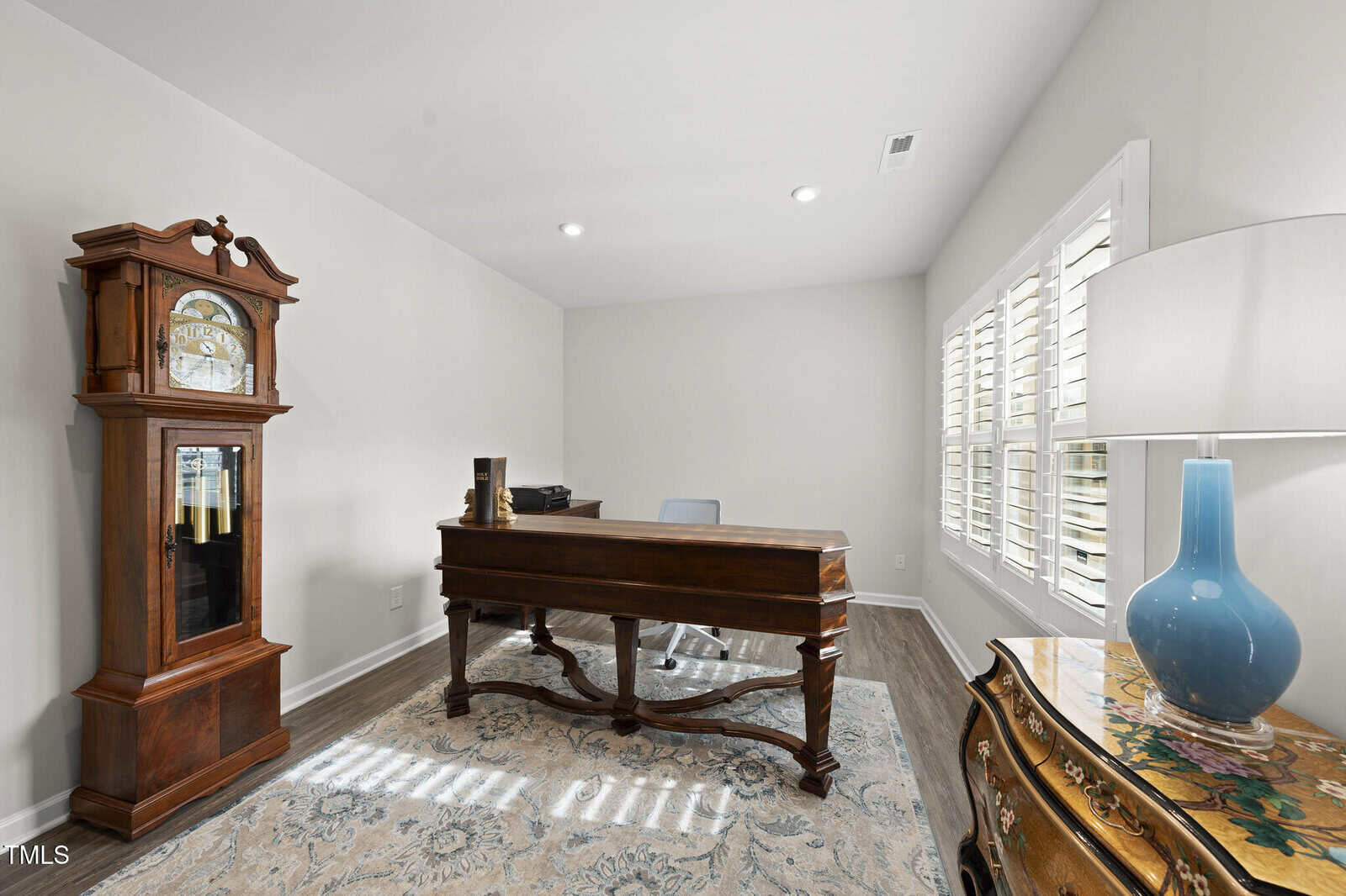 A home office with a wooden desk, blue chair, grandfather clock, ornate side table with a blue lamp, and a large window with white shutters. The room has light walls and a patterned rug on the floor.