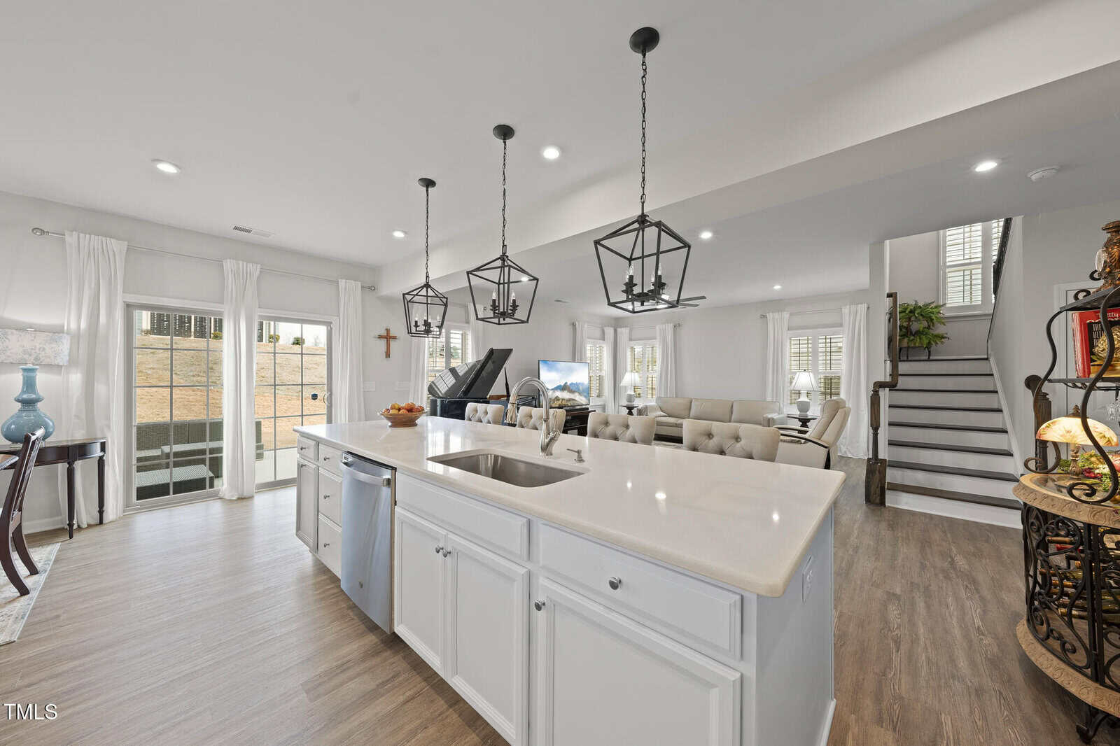 Modern open-concept living area with a kitchen island, white cabinetry, stainless steel appliances, pendant lights, dining space, and a living room. Stairs lead up, and large windows provide ample natural light.