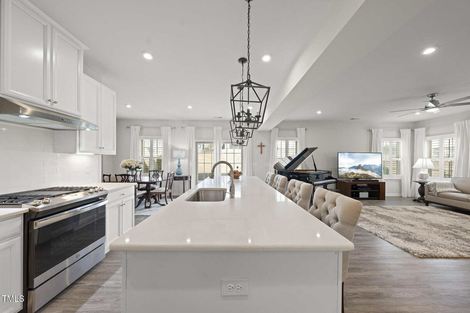Modern open-concept kitchen and living area with white cabinets, stainless steel appliances, an island with barstools, a grand piano, a TV, and a beige sofa on a large rug. Natural light fills the space through many windows.
