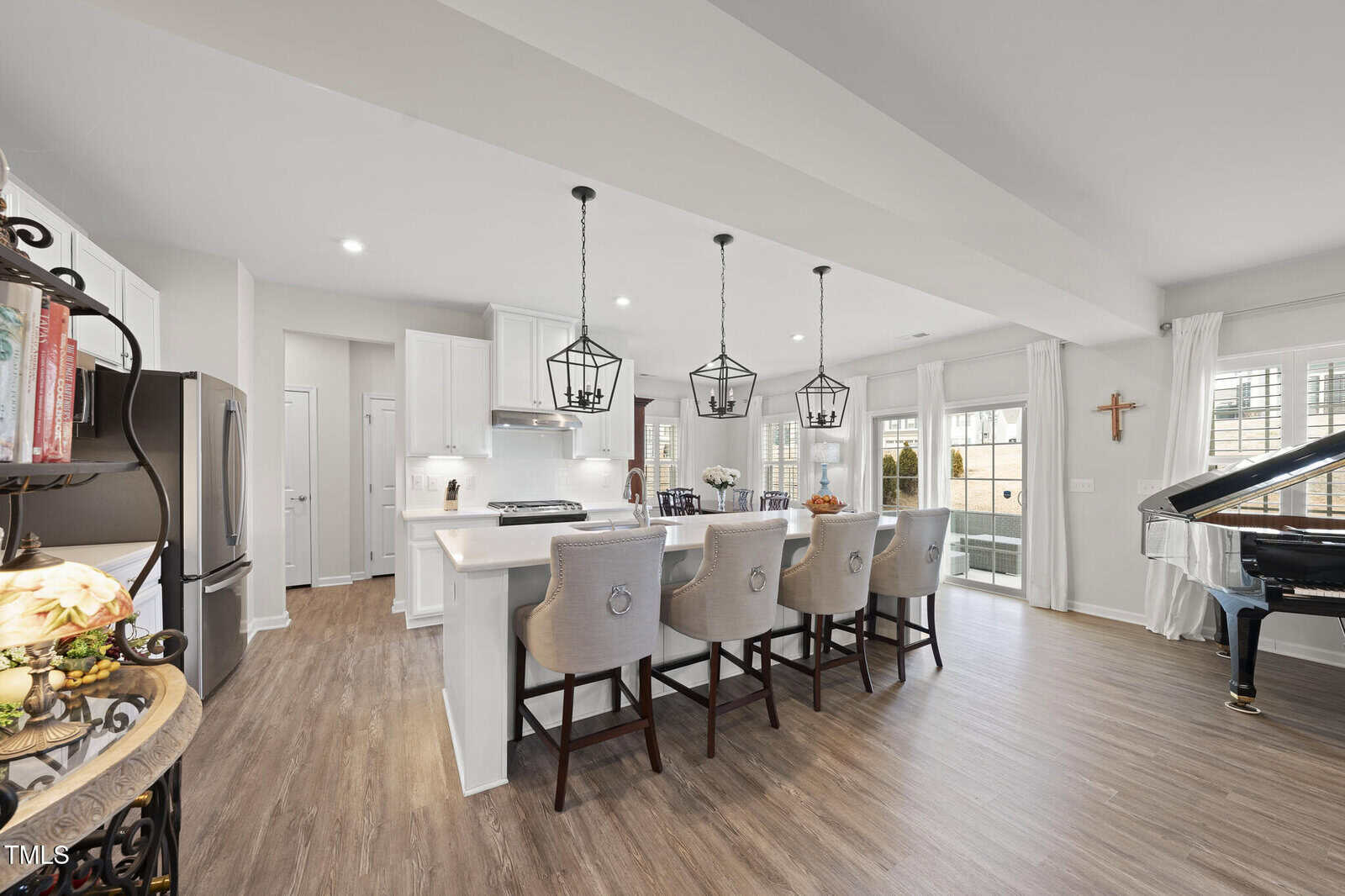 A modern kitchen with white cabinets, a large island with four beige chairs, three pendant lights above, stainless steel appliances, and a grand piano near windows with white curtains in the background.