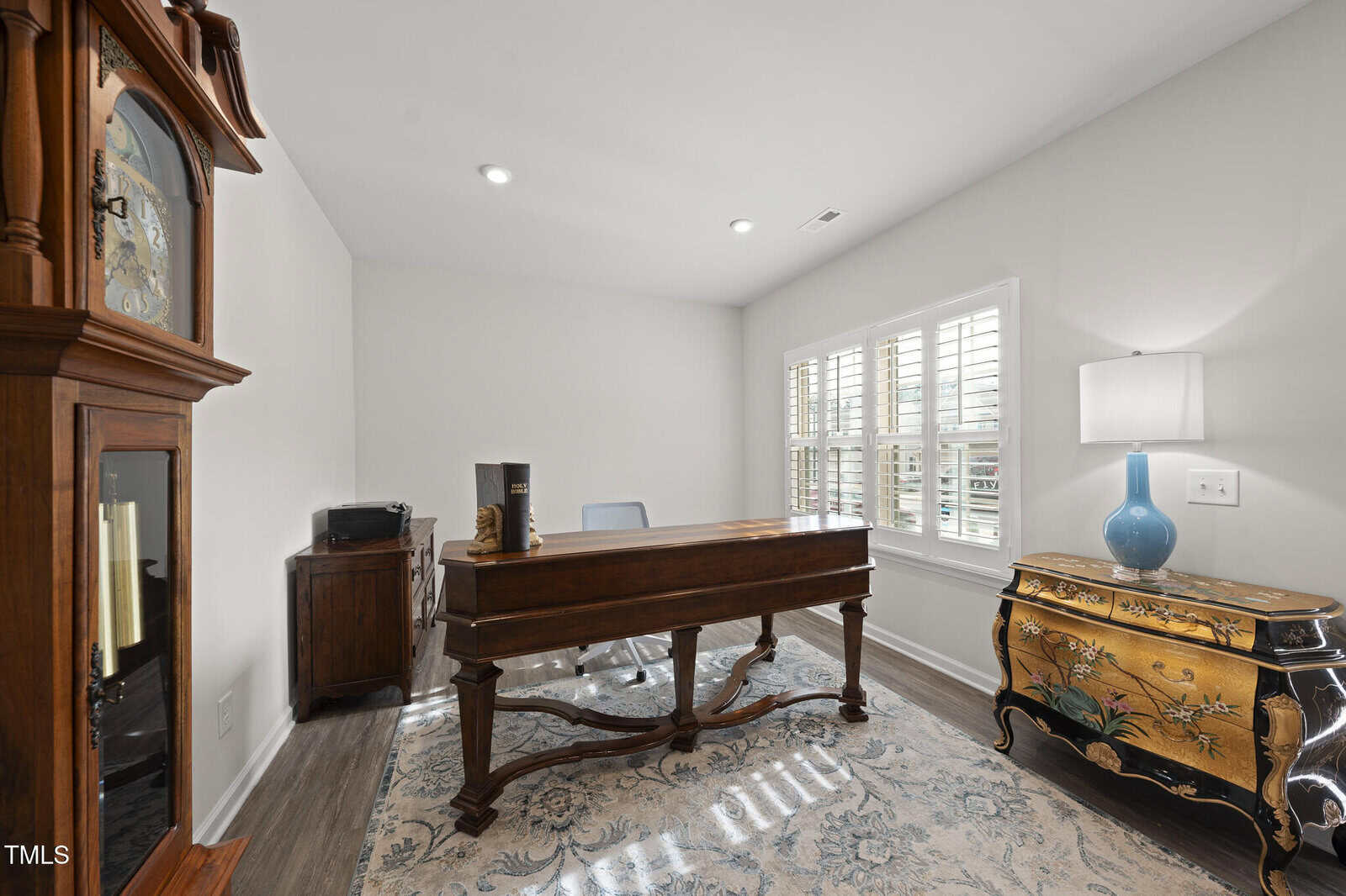 A home office with a wooden desk, a blue and gold chest, a grandfather clock, a lamp with a blue base, an office chair, a printer, and a large window with white shutters letting in natural light.