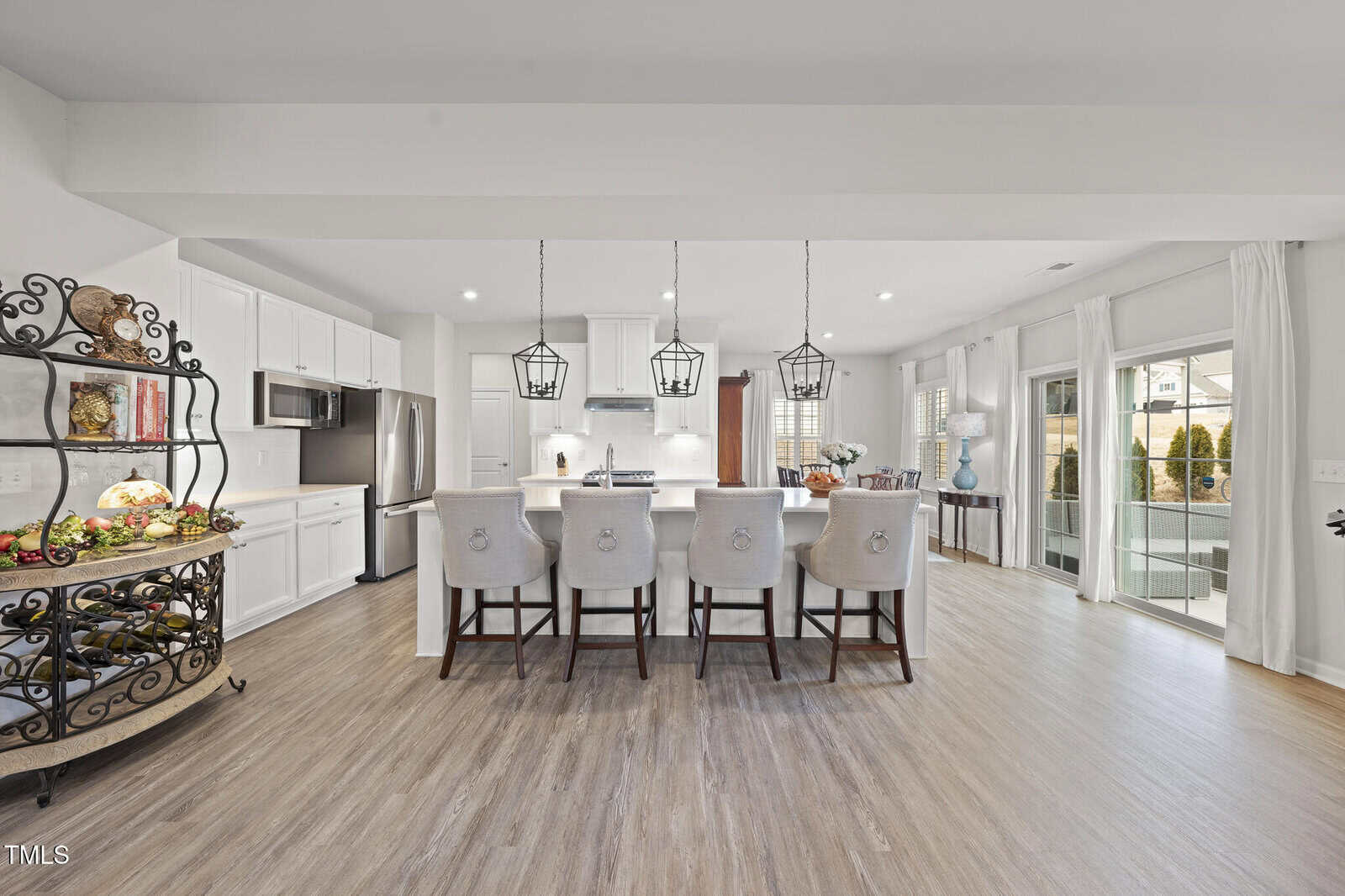 Modern kitchen with white cabinets, stainless steel appliances, island with four upholstered chairs, pendant lights, open shelving, and sliding glass doors leading to a patio; wood flooring throughout the space.