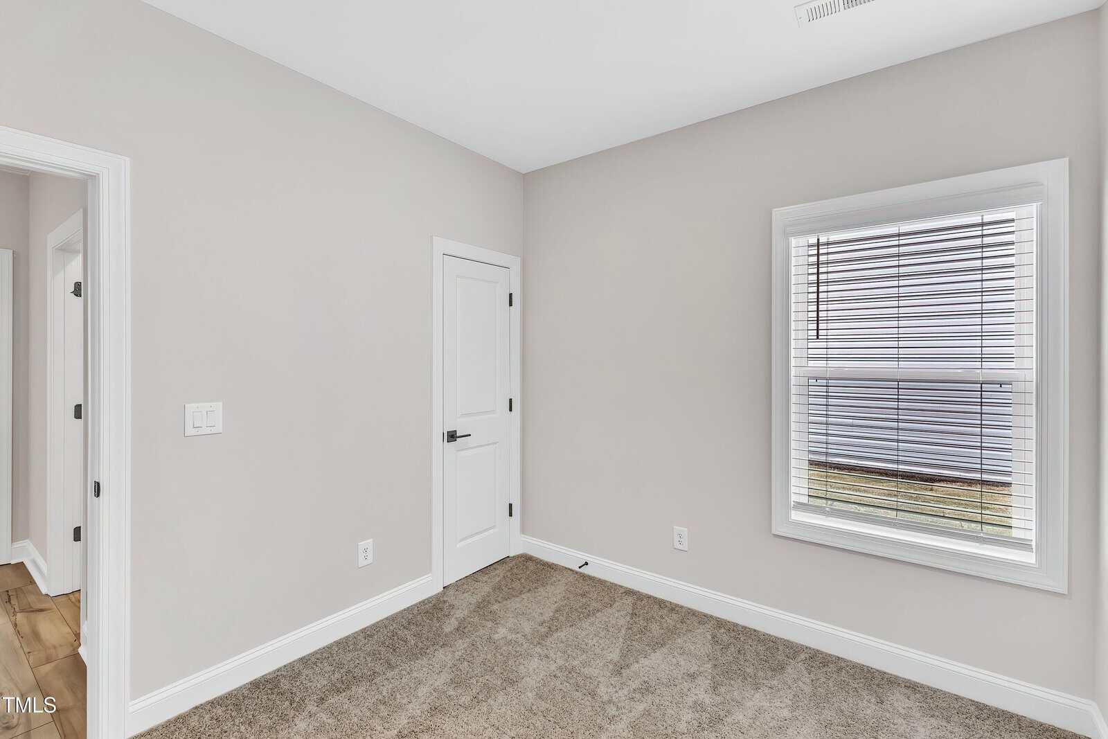 A small empty room with beige carpet, light gray walls, a white door, and a window with closed blinds. Natural light enters through the window, and hardwood flooring is visible in the adjacent hallway.