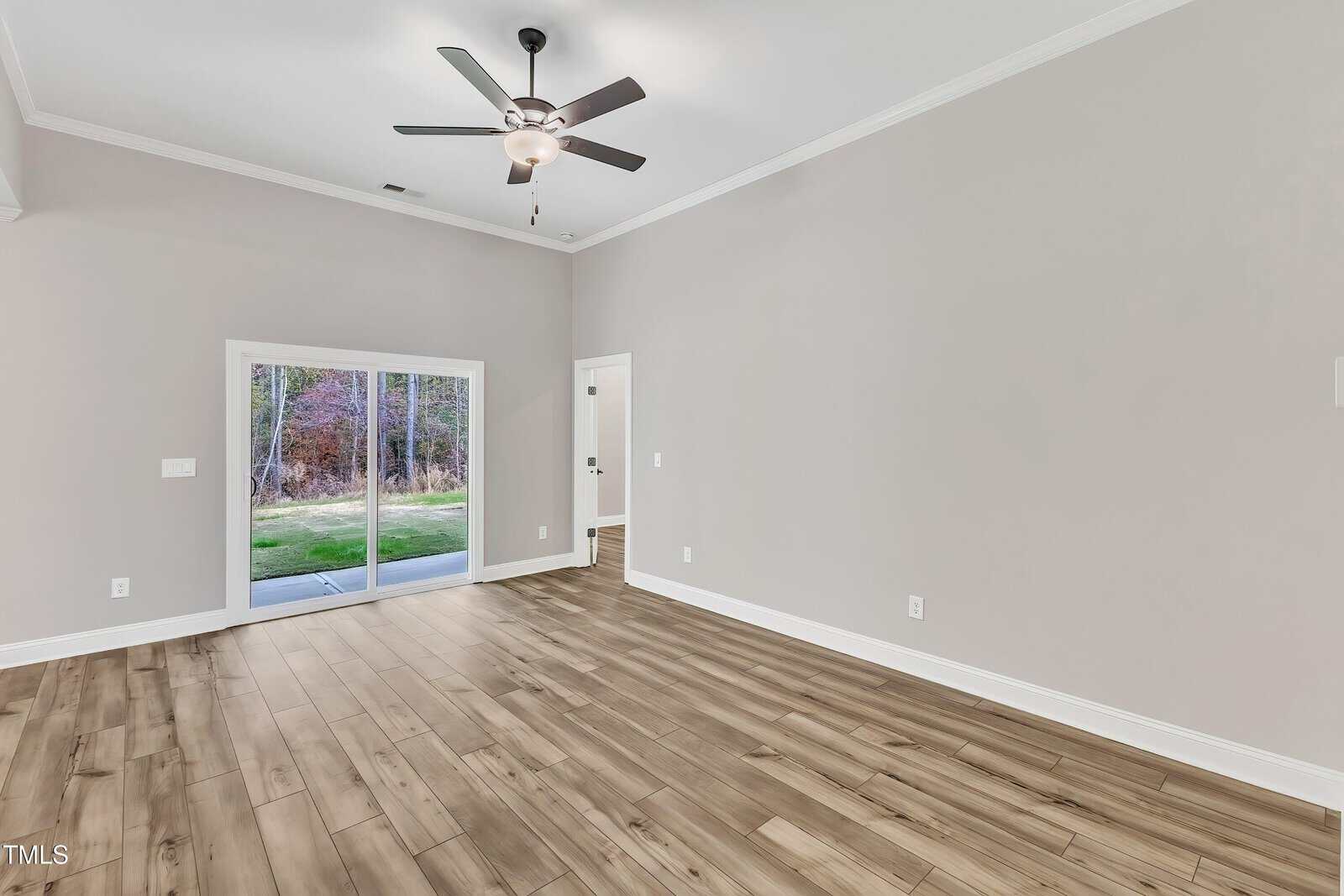 A spacious, empty room with light wood flooring, light gray walls, a ceiling fan with lights, and a large sliding glass door leading outside. Natural light enters through the door, and there are several outlets on the walls.