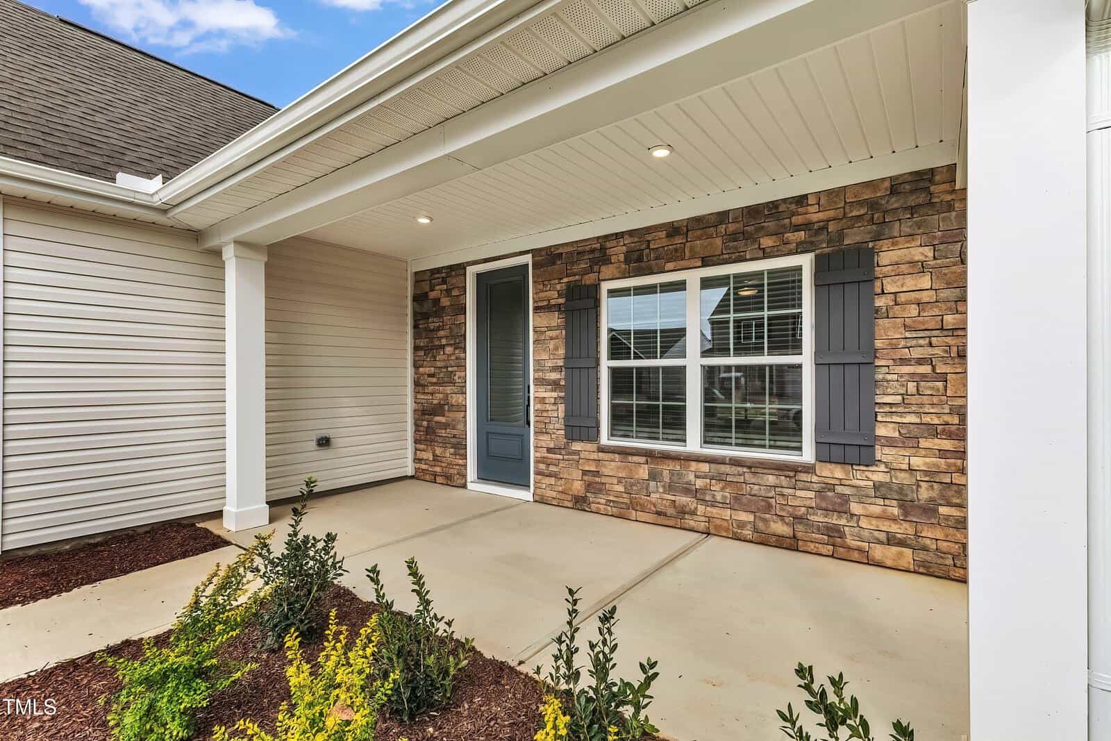 A covered front porch with a concrete floor, stone accent wall, blue front door, and a large window with gray shutters. Small shrubs and plants border the edge of the porch.