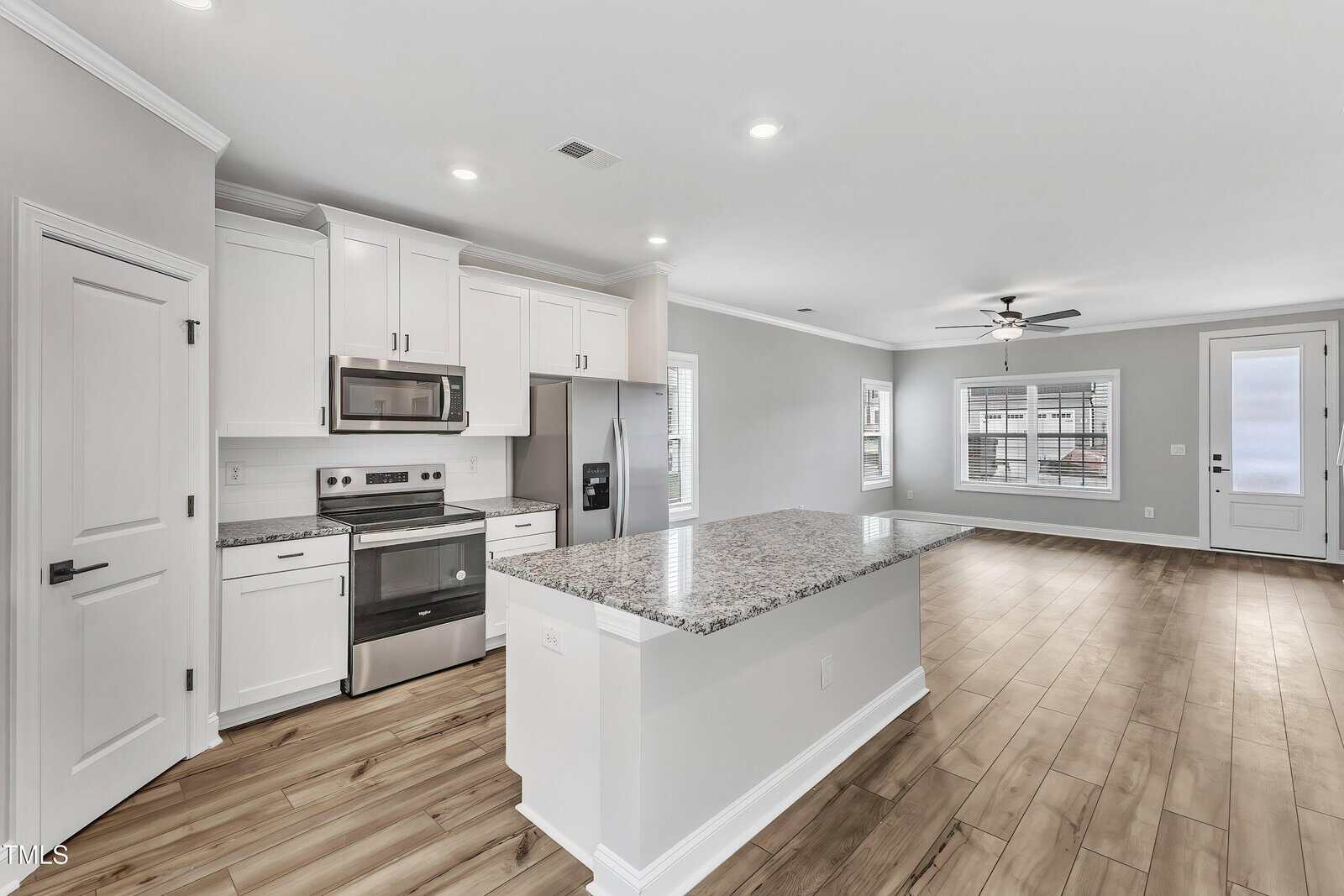 Modern kitchen with white cabinets, stainless steel appliances, granite island, and wood flooring, open to a bright living area with windows, ceiling fan, and glass door to the outside.