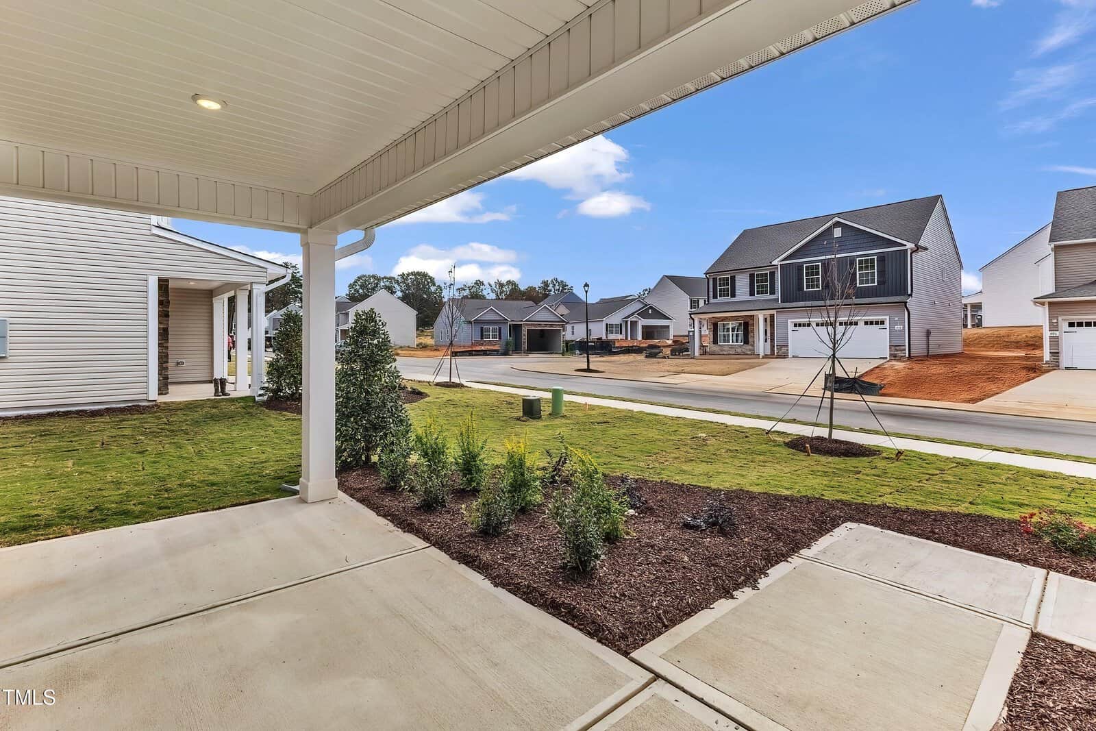 View from a covered porch looking out onto a suburban street with several newly built houses, fresh landscaping, and a lawn with young plants and trees. Concrete sidewalk and driveway in the foreground.