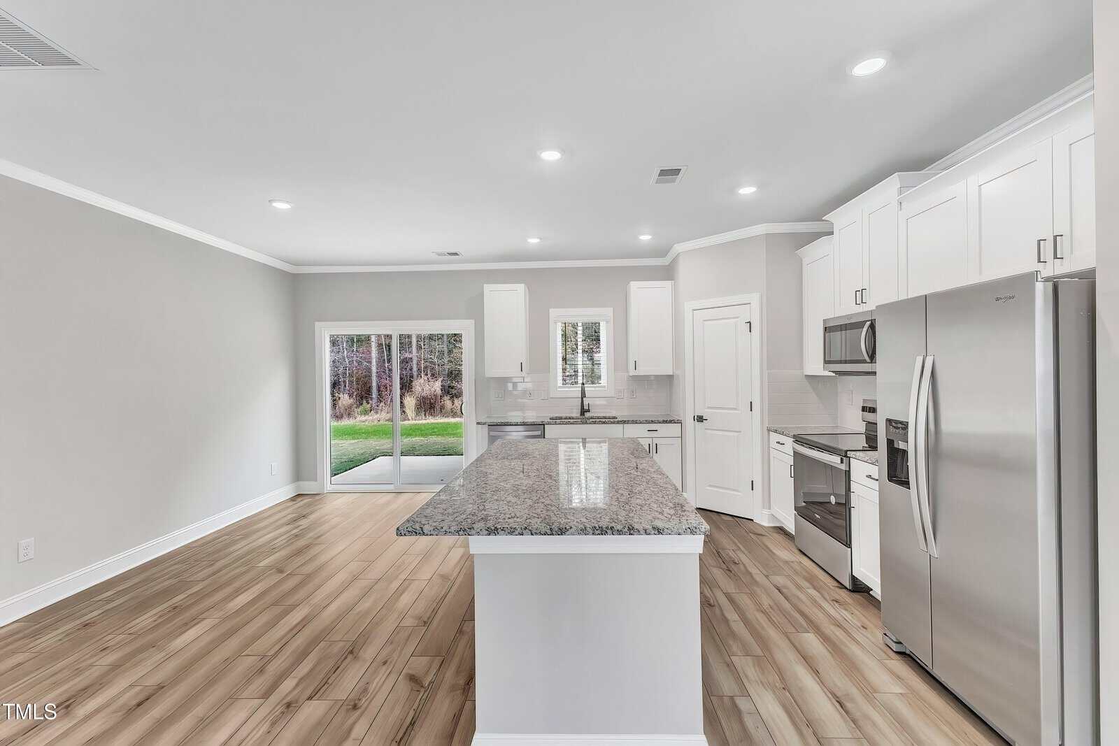 Modern kitchen with white cabinets, stainless steel appliances, granite island, wood-look flooring, and sliding glass doors leading to a backyard; neutral walls and recessed lighting throughout the space.