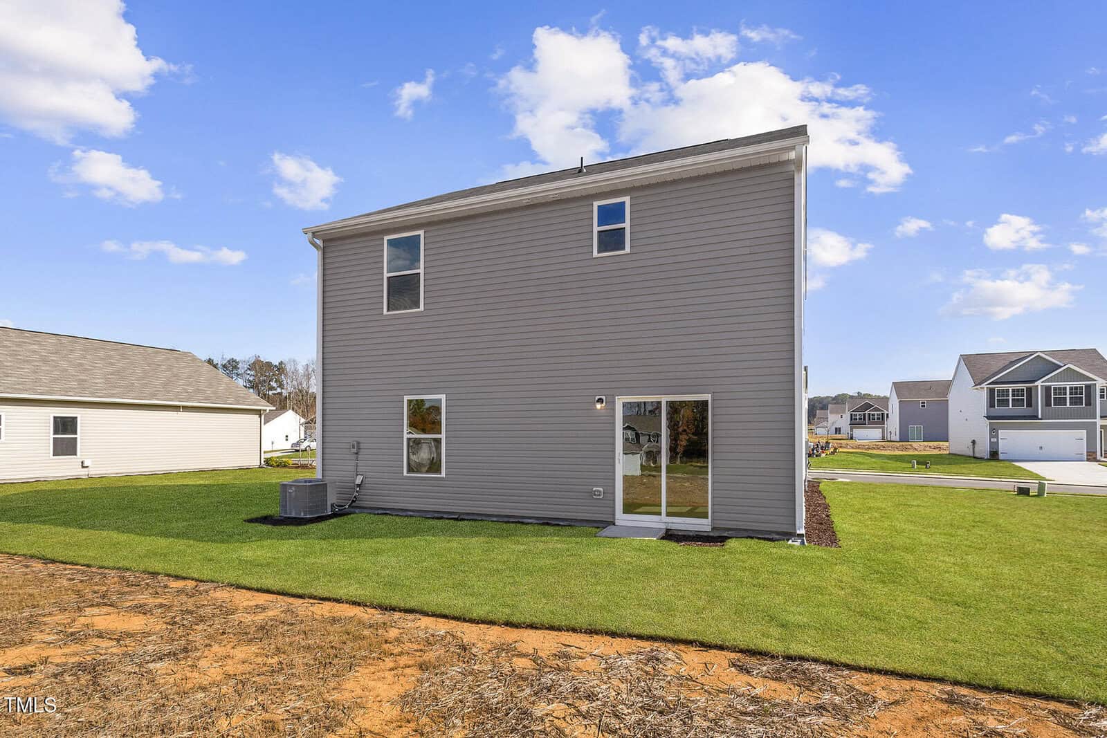 A two-story gray house with a few small windows and glass patio door stands in a grassy backyard. Adjacent houses are visible in the background under a clear blue sky with scattered clouds.