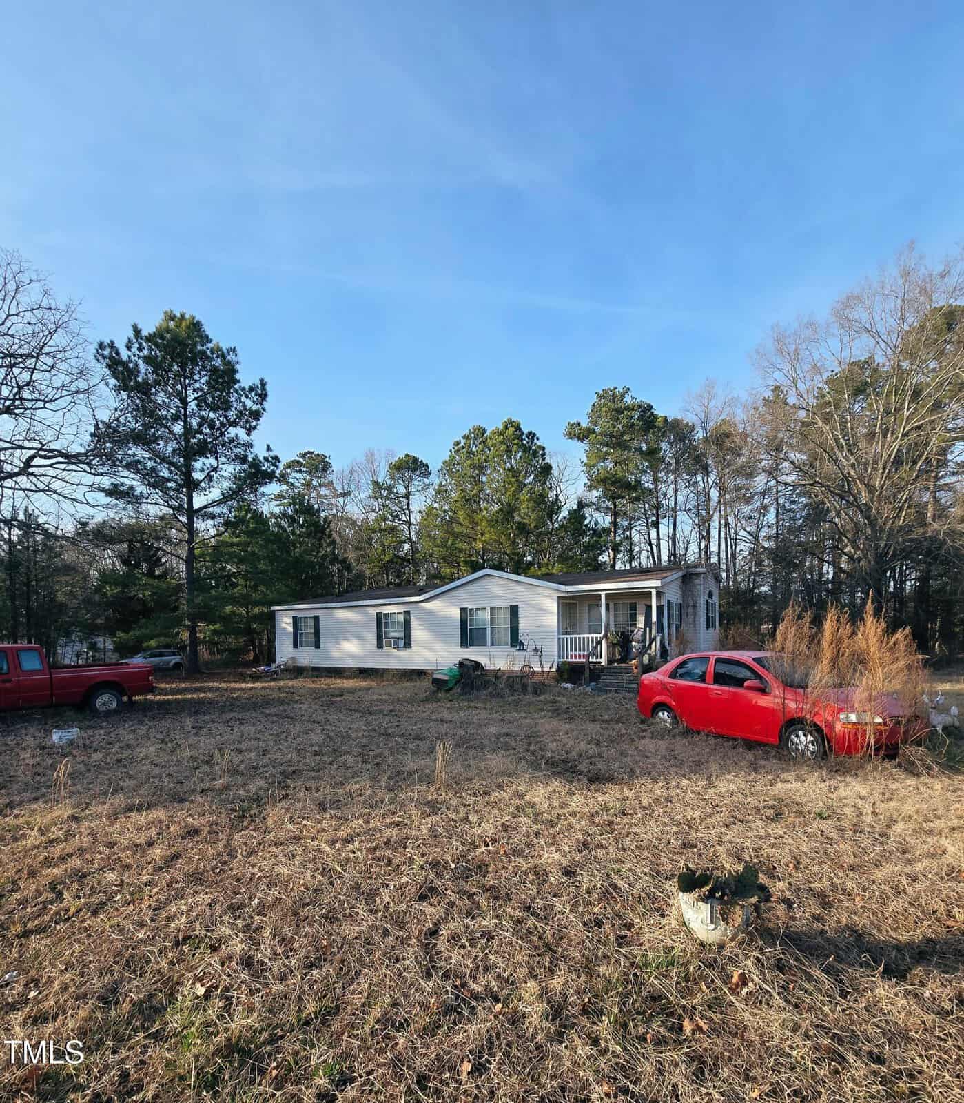 A small, single-story house at 2089 Lisa Lane in Creedmoor features white siding and sits in a grassy yard with several trees. A red car and truck are parked in front. The porch, adorned with chairs, offers a relaxing spot under the clear sky.