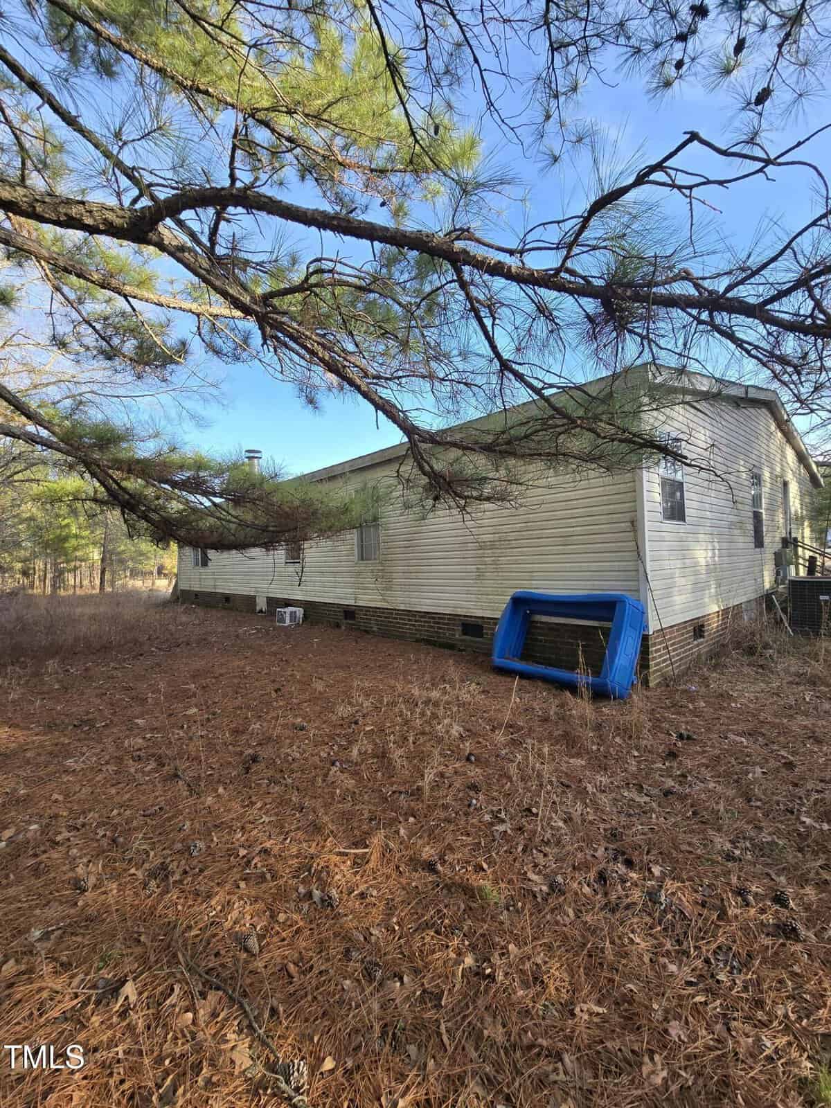 A single-story mobile home with beige siding and a green roof is surrounded by pine trees. An upside-down blue slide lies next to the house. The ground is covered with pine needles. The sky is clear and sunny.