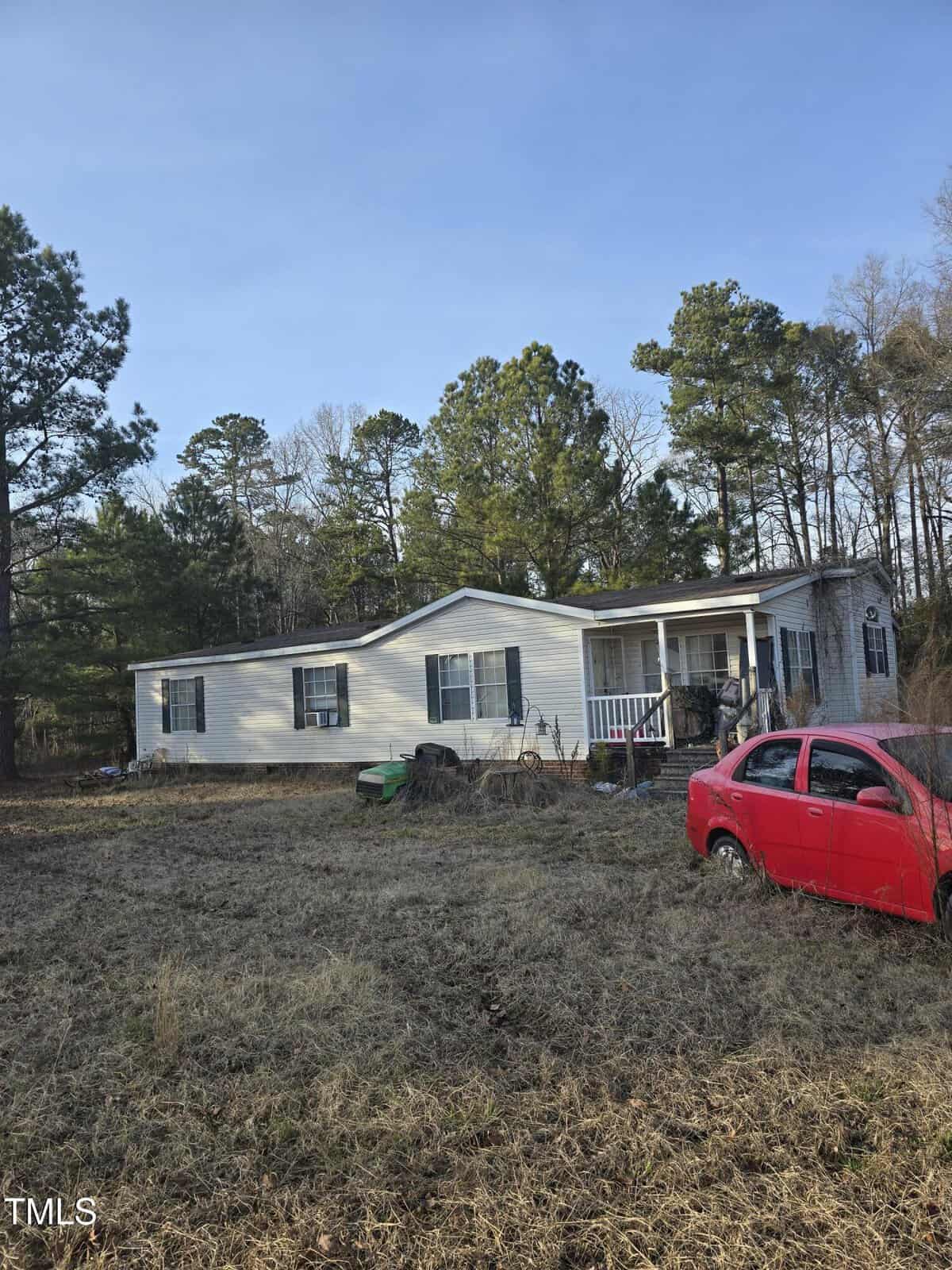 A single-story manufactured home with light siding and dark shutters stands in a grass-covered yard. A small porch is at the entrance. A red car is parked on the right. Trees surround the property under a clear sky.