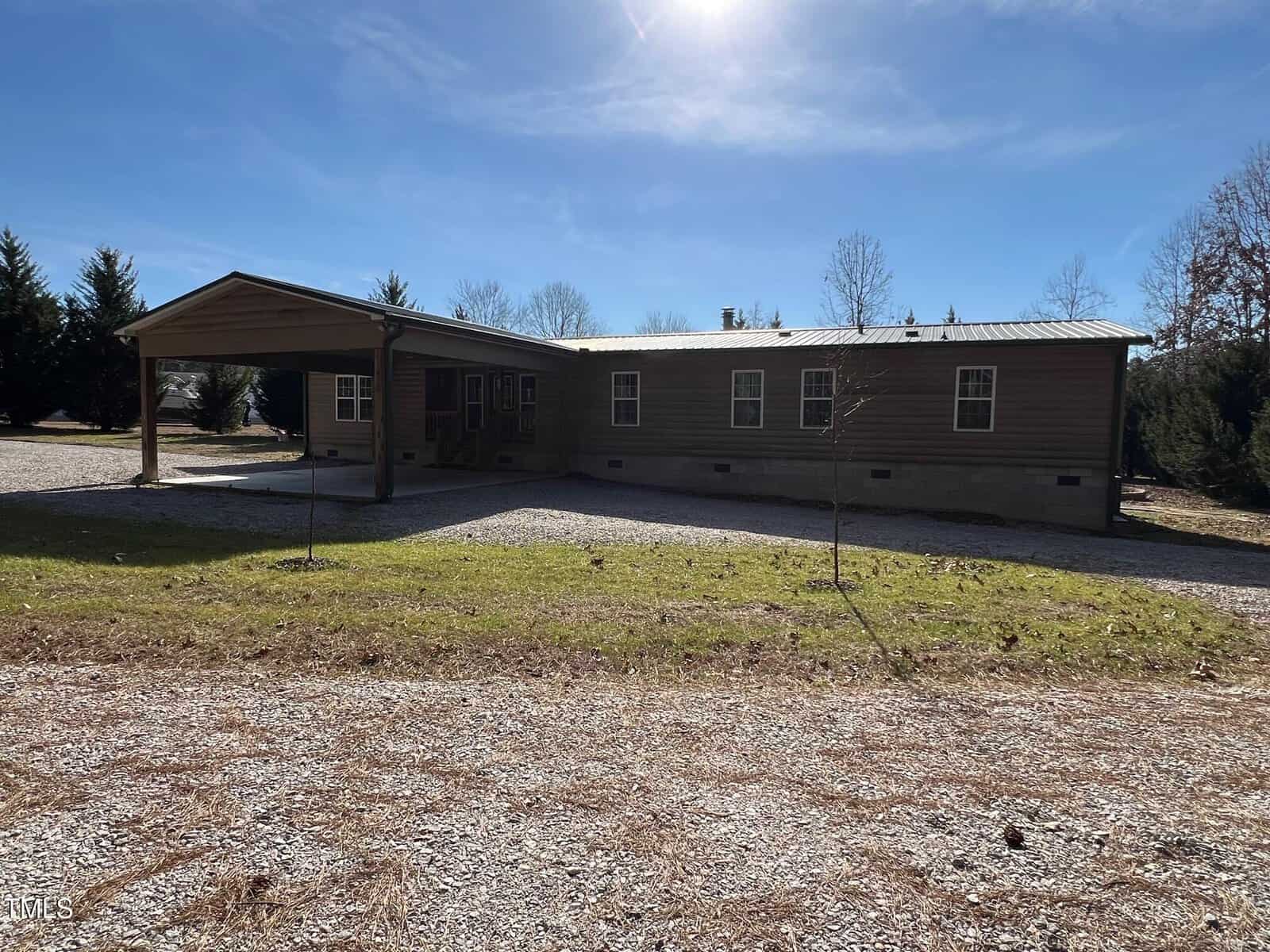 A single-story, rectangular wooden house with a metal roof is shown. It has a covered carport on the left side. The surrounding area is grassy with trees in the background. The sky is clear and sunny.