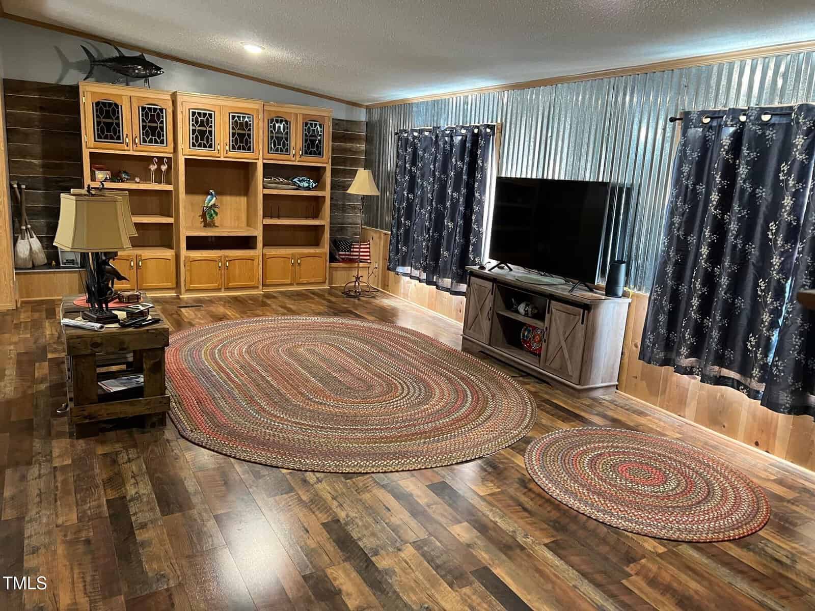 Living room with wooden flooring and walls. A large braided oval rug and a smaller matching rug on the floor. Entertainment center with a TV, bookshelves with decorative items, side table with a lamp, and star-patterned curtains on the windows.