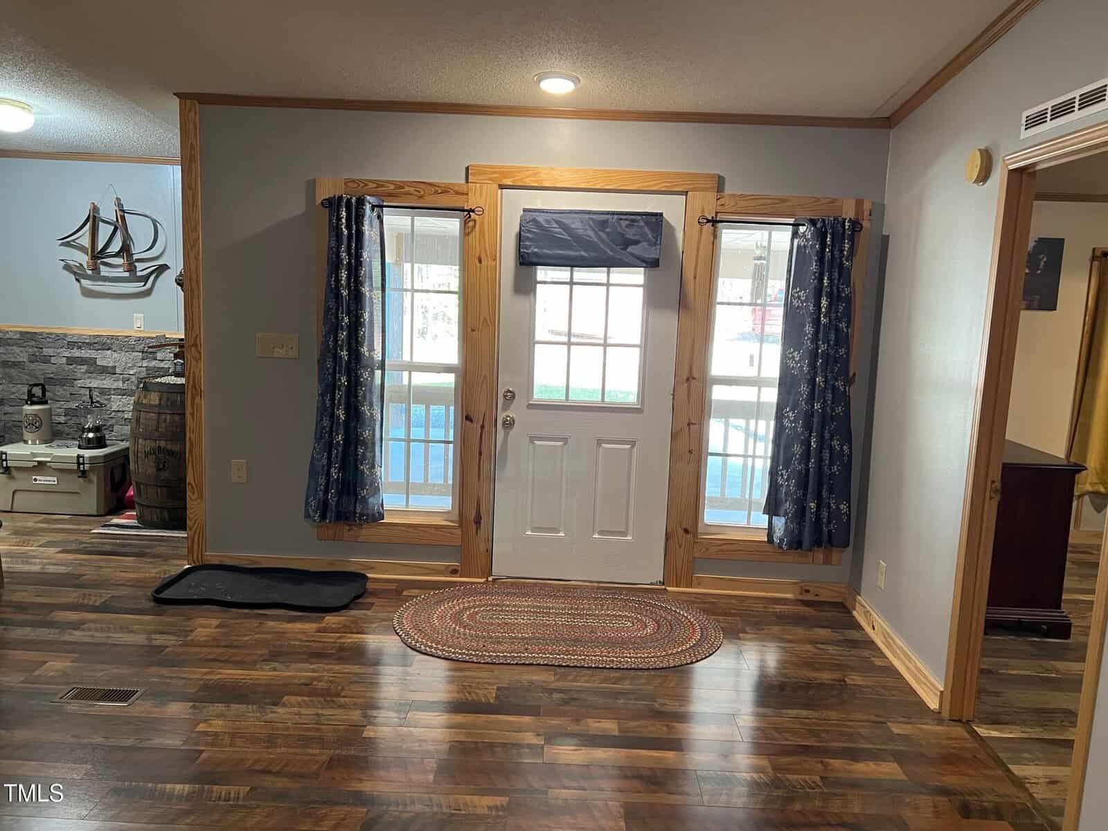 A room with a wooden floor features a white door with a window at the top, flanked by two windows covered with dark curtains. A round rug lies in front of the door. A decorative ship and a barrel are visible to the left.
