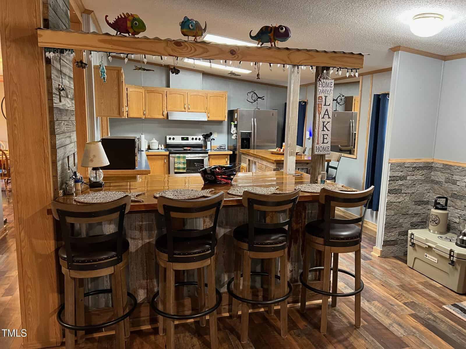 A cozy kitchen and bar area with wooden cabinets and countertops. Four bar stools line the counter. The room features hardwood floors and stone accents. Decorative items, like colorful lizard figurines, are placed above the bar.